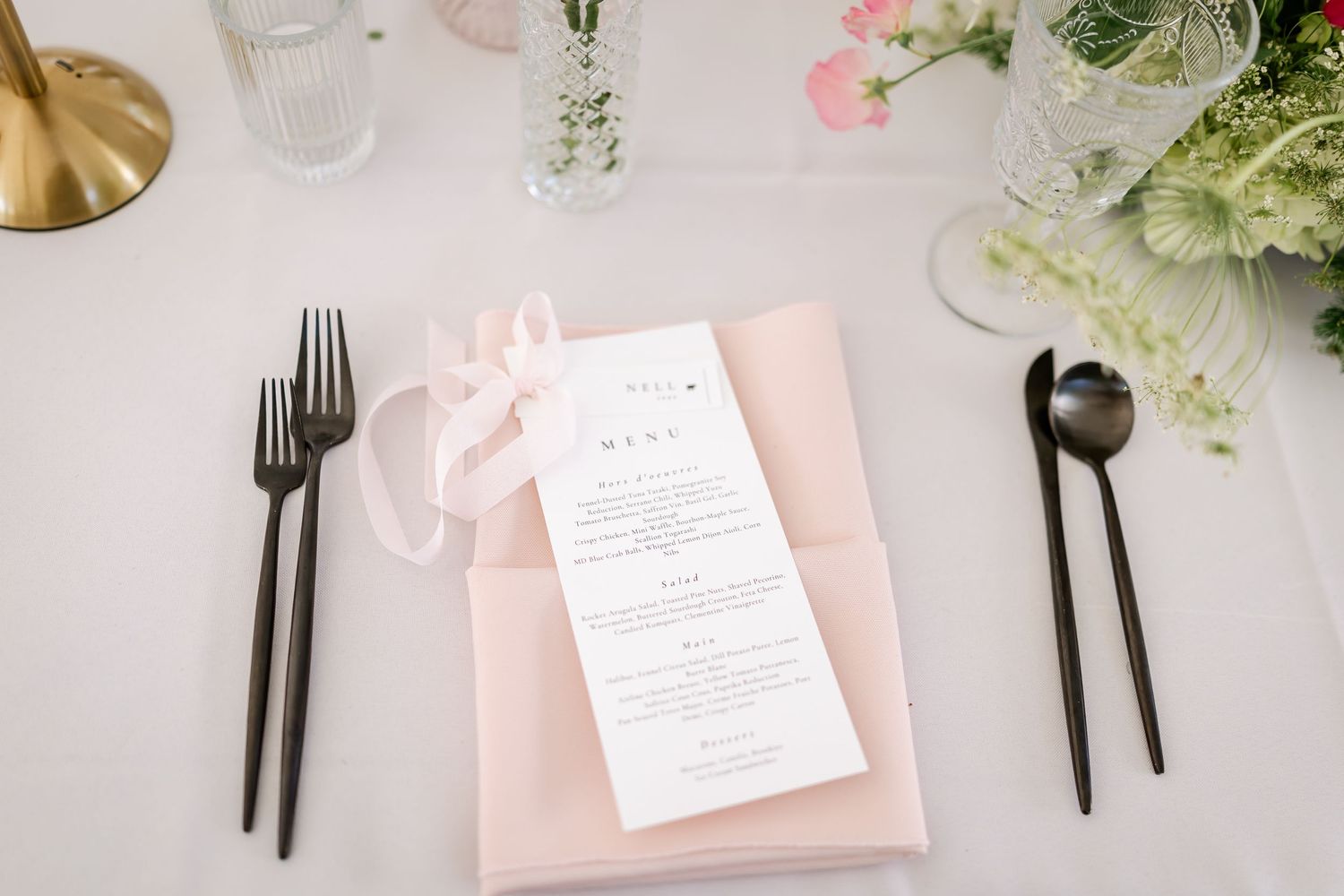 Elegant place setting with pink napkins, menu cards and modern black flatware on white tablecloth.