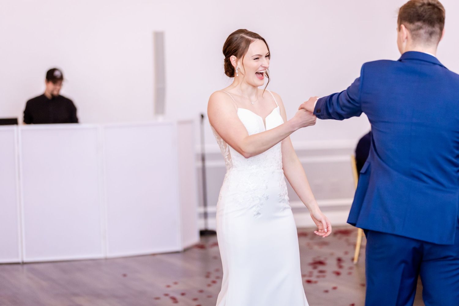 A bride in a white dress laughs and dances with a partner in a blue suit at a wedding celebration.