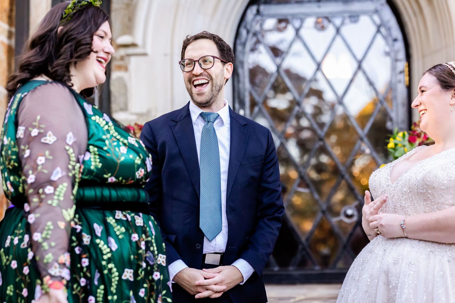 Three wedding guests share a candid laughing moment in front of a decorative glass window.