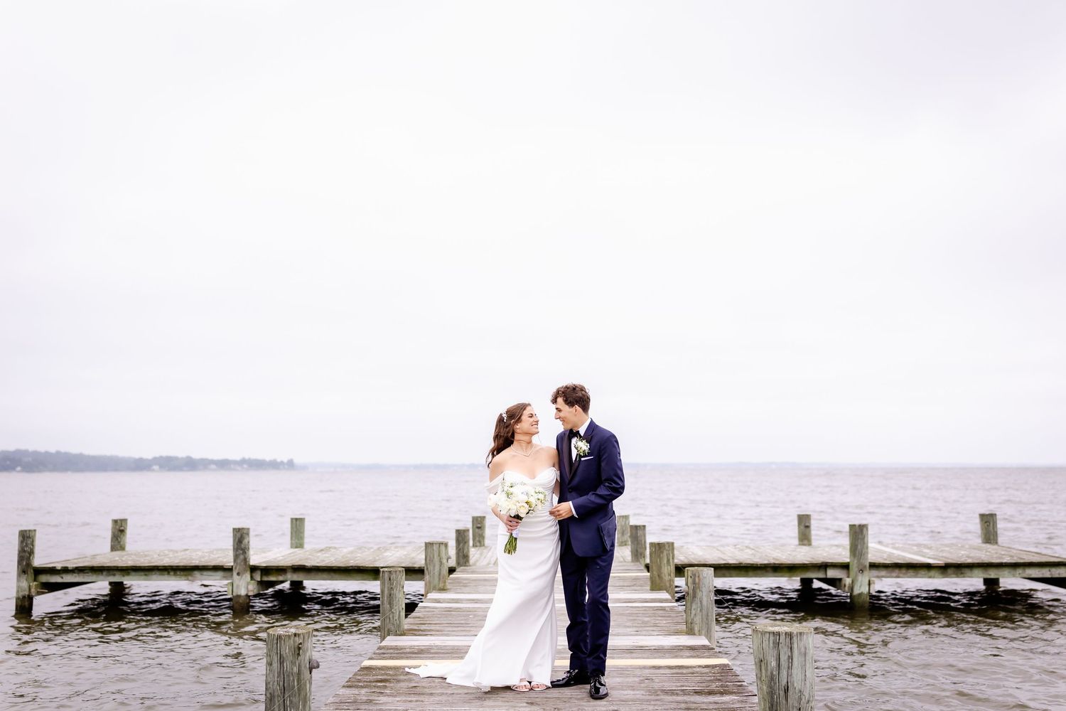 A couple in wedding attire embraces on a wooden pier overlooking a misty waterfront on their special day.