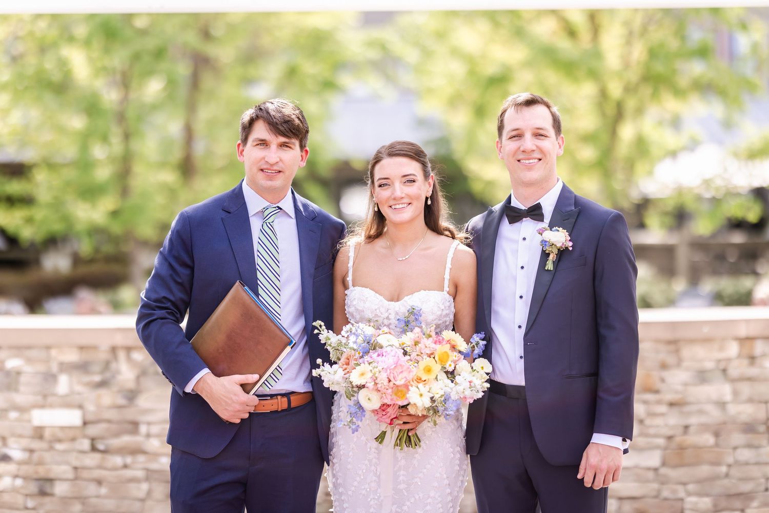 Three people pose formally outdoors against stone wall backdrop.