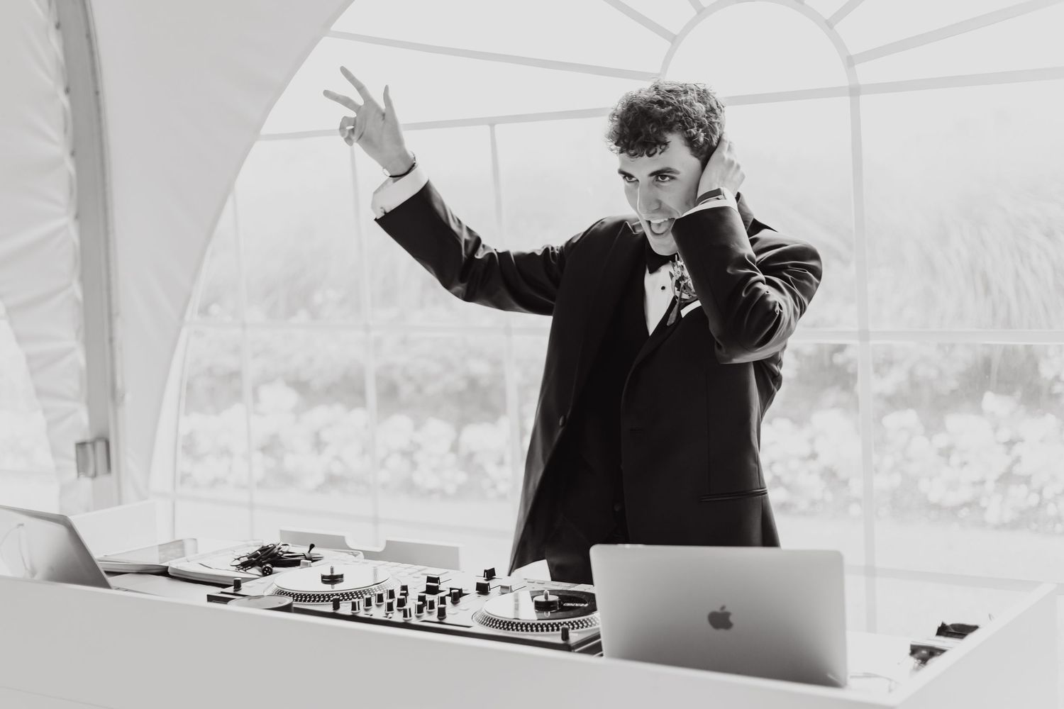 Wedding DJ plays music from behind a professional setup with equipment and laptop at an outdoor reception.