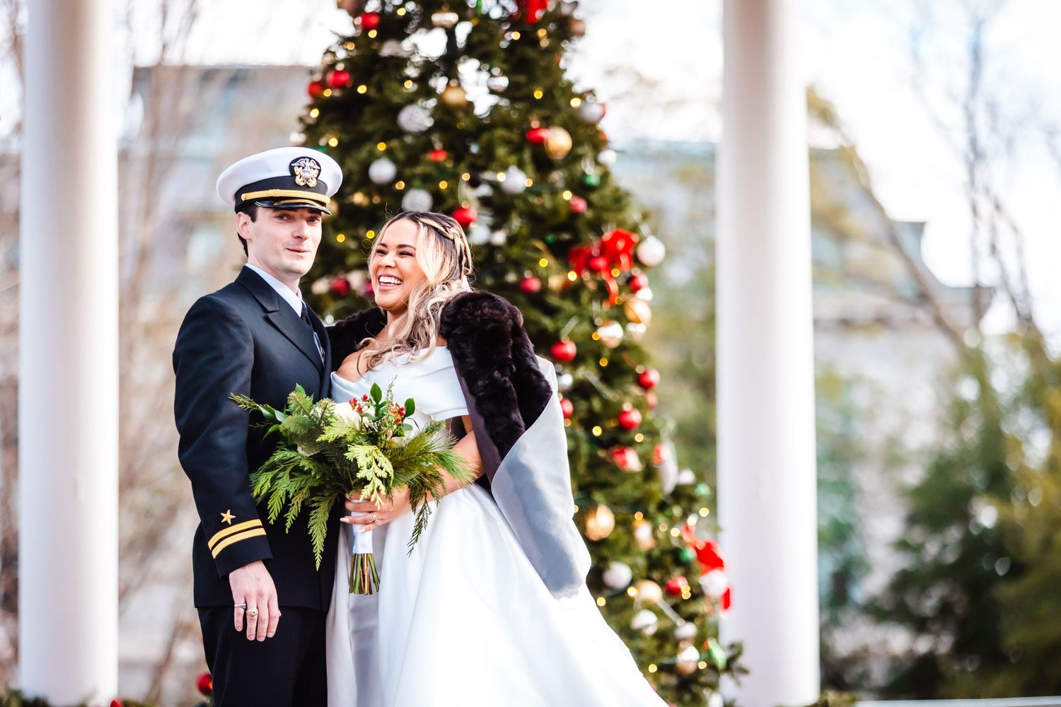 A military wedding couple shares a romantic moment in front of a decorated Christmas tree with white columns.