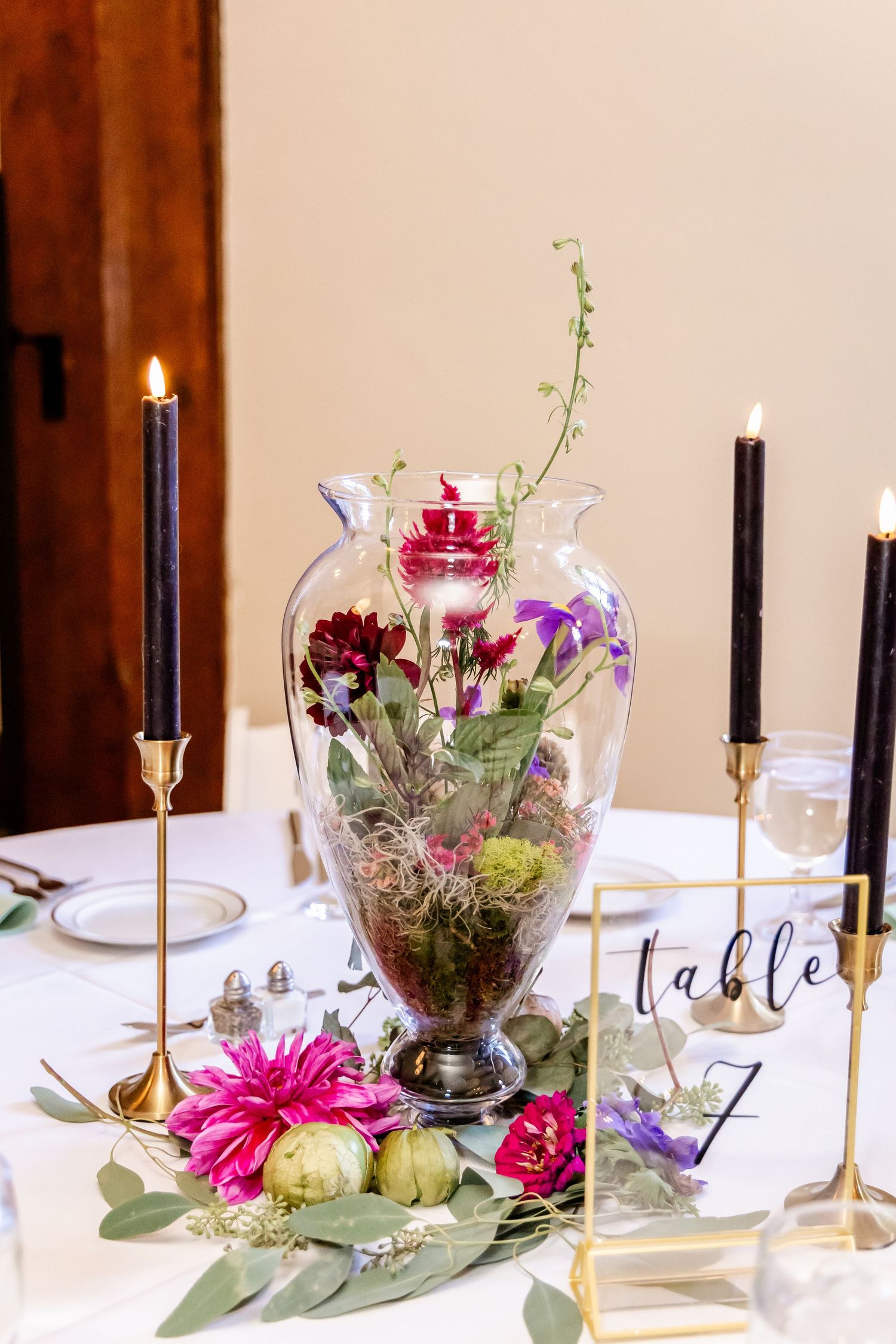 Elegant wedding table centerpiece featuring a glass vase with colorful flowers and tall black candles in gold holders.