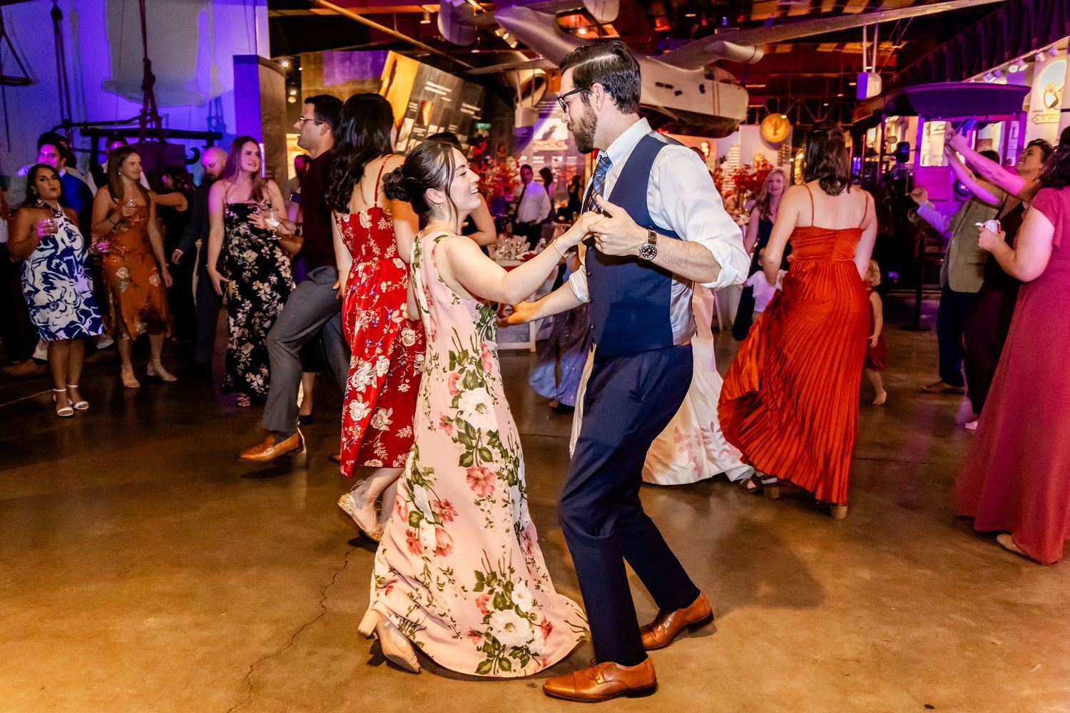 Wedding guests dance together on a rustic venue dance floor with purple mood lighting.