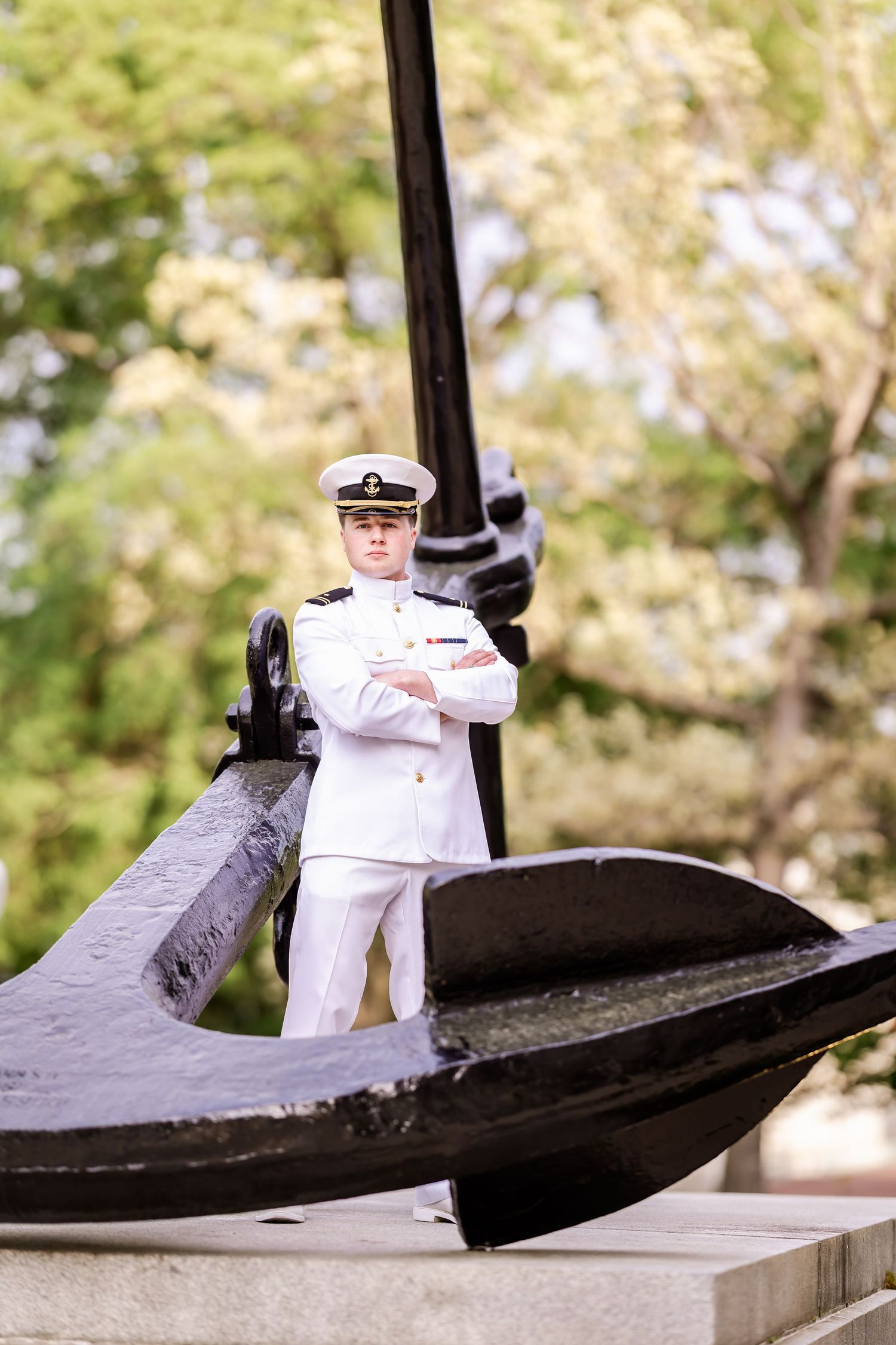 Naval officer in white uniform poses with crossed arms beside black anchor monument.