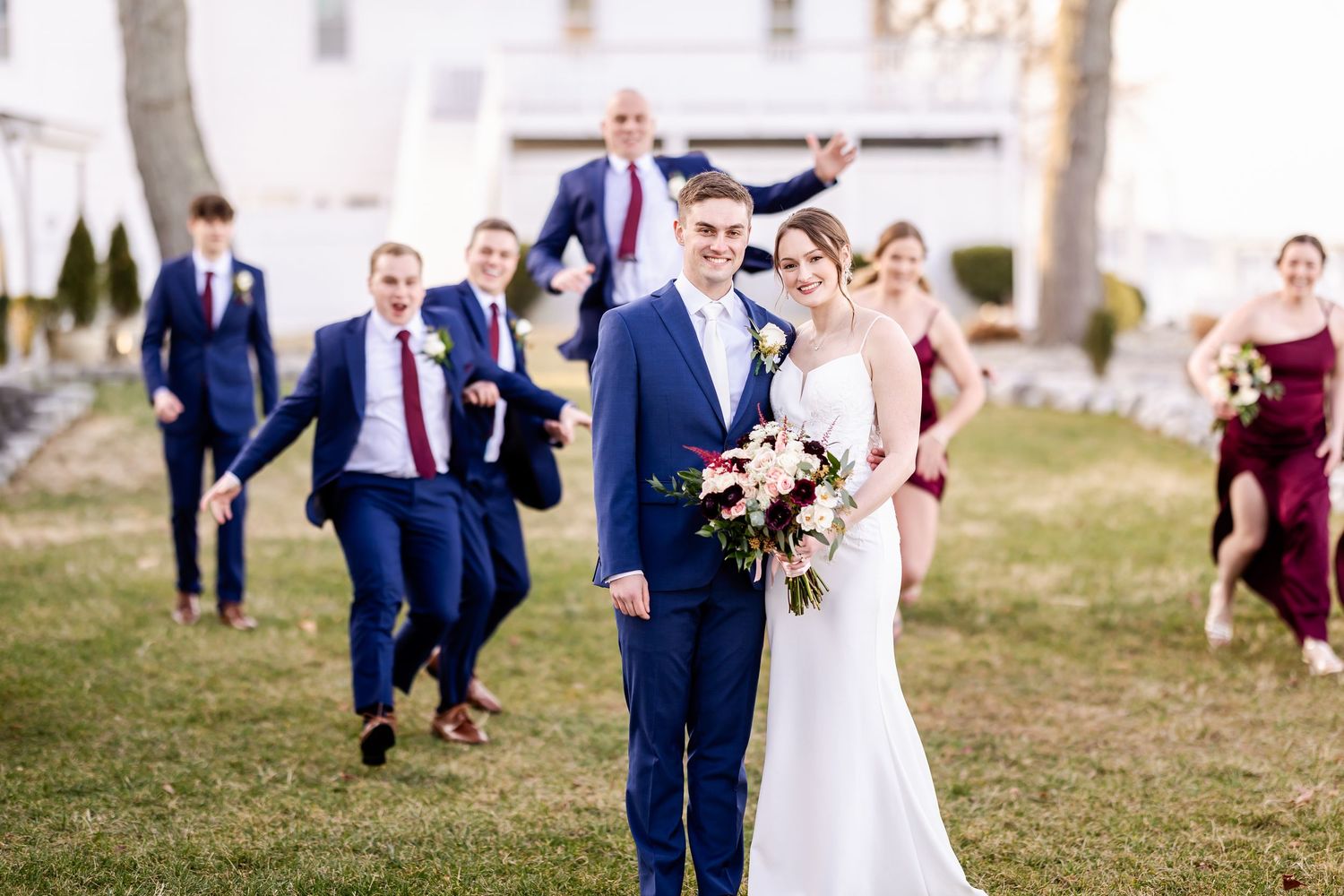 Wedding party in navy suits and burgundy dresses poses outdoors for group photos on a sunny autumn day.