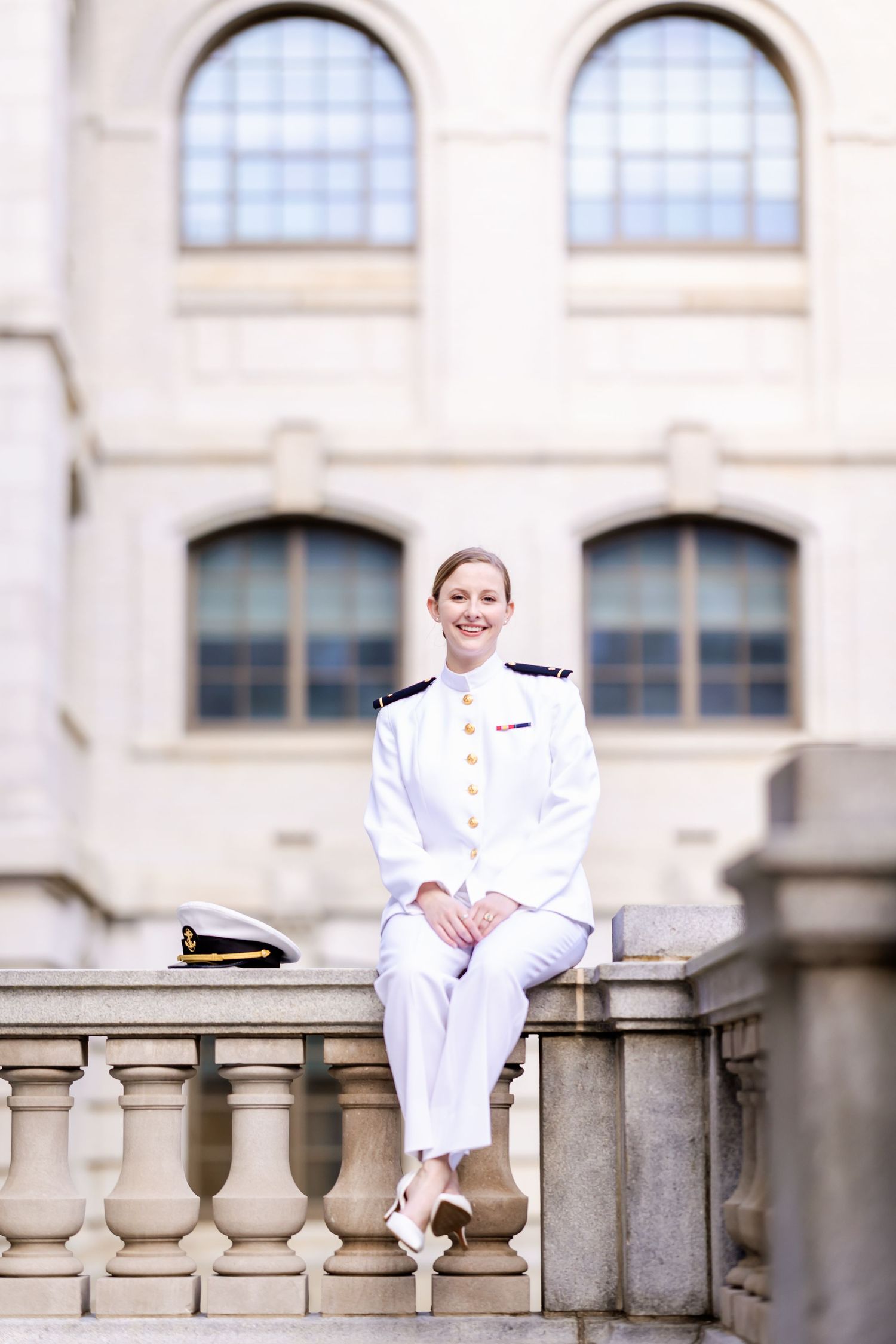 Officer in white naval uniform sits gracefully on stone balustrade in front of arched windows.