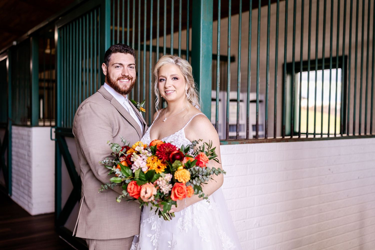 A bride and groom pose together at their wedding, with bright orange and red floral bouquets and beige attire.