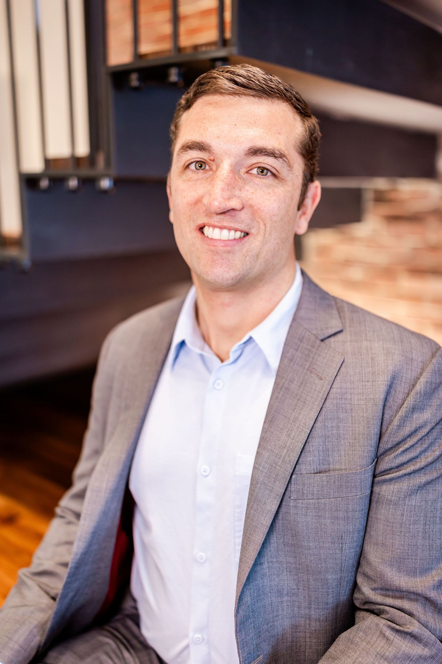Professional headshot of person in grey suit jacket and light blue shirt against wooden backdrop.