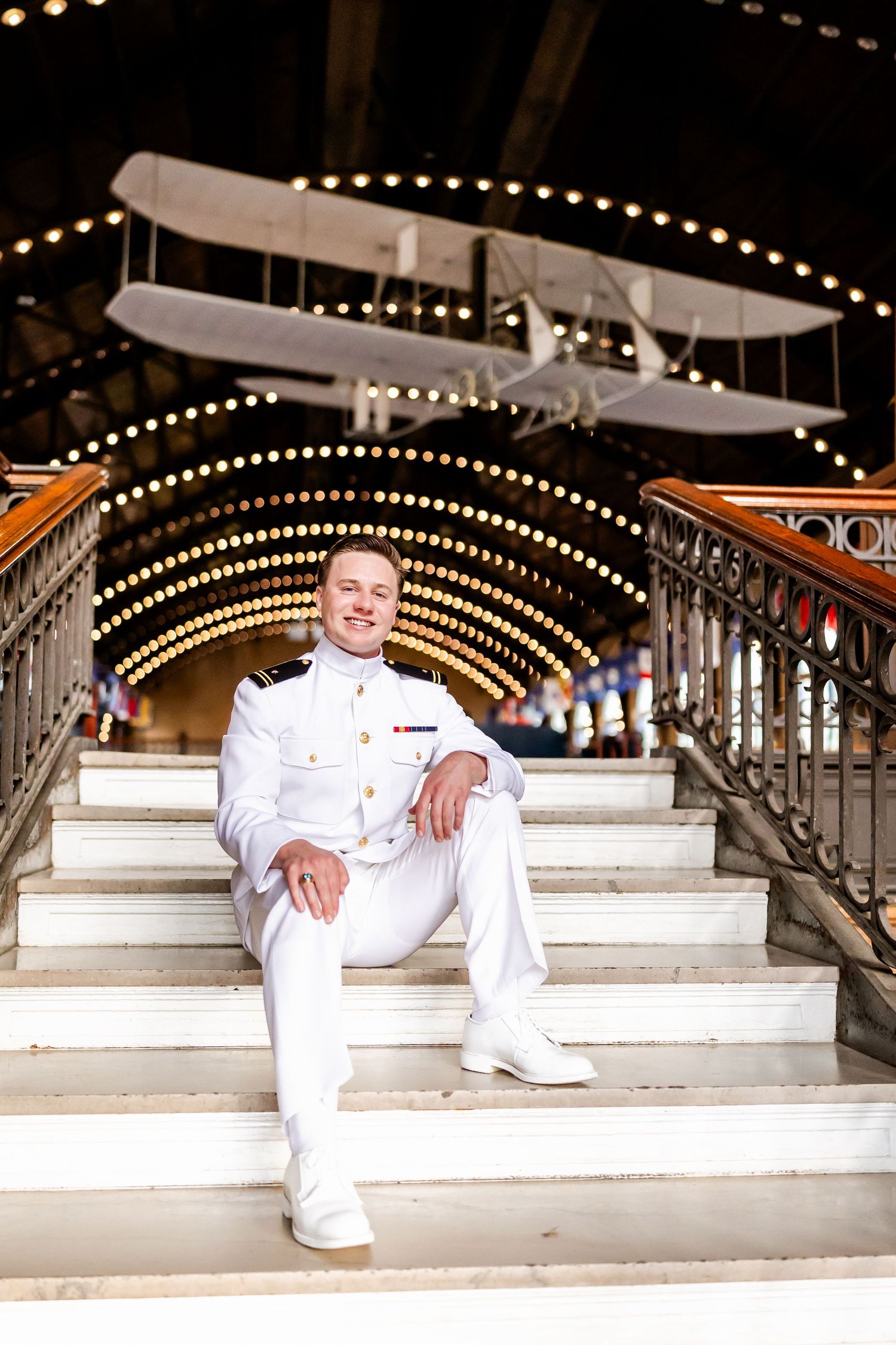 Military officer in white uniform relaxing on ornate steps with string lights in background.