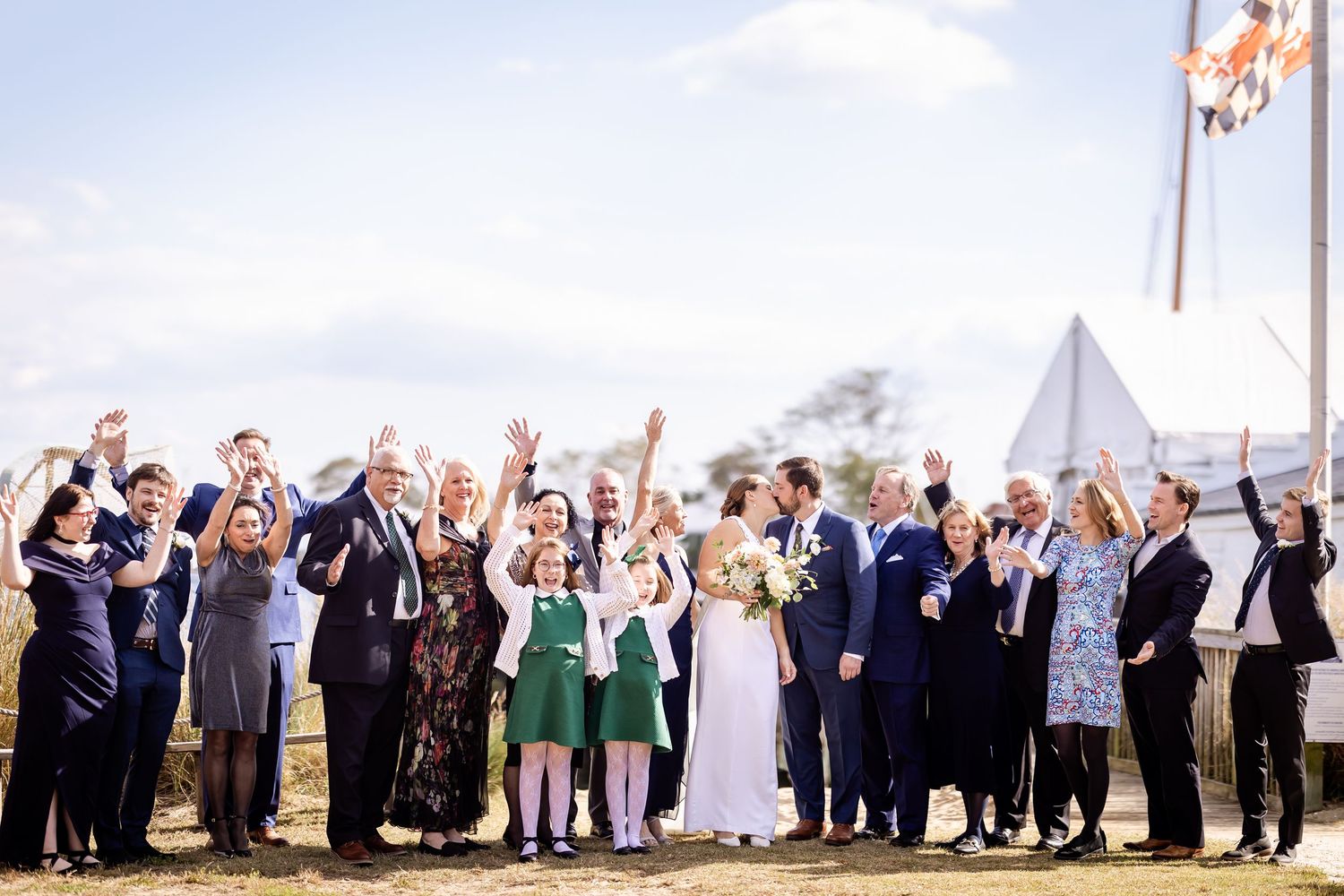 Large wedding party celebrates outdoors with raised arms near white tent venue.