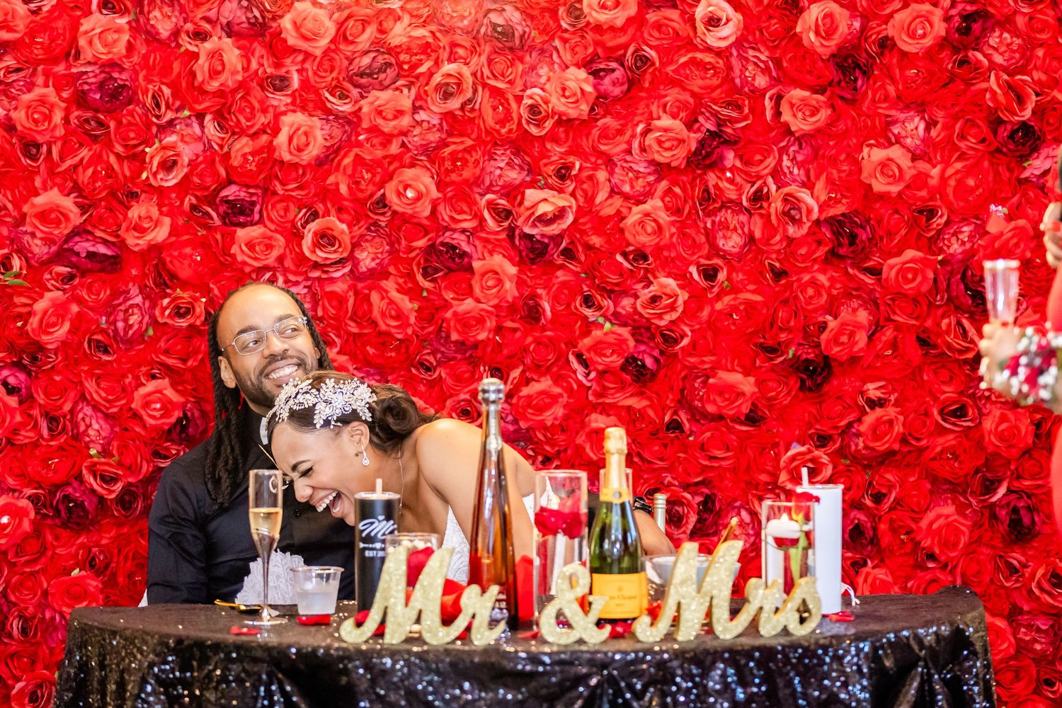 Wedding reception table decorated with champagne bottles against vibrant red rose wall.