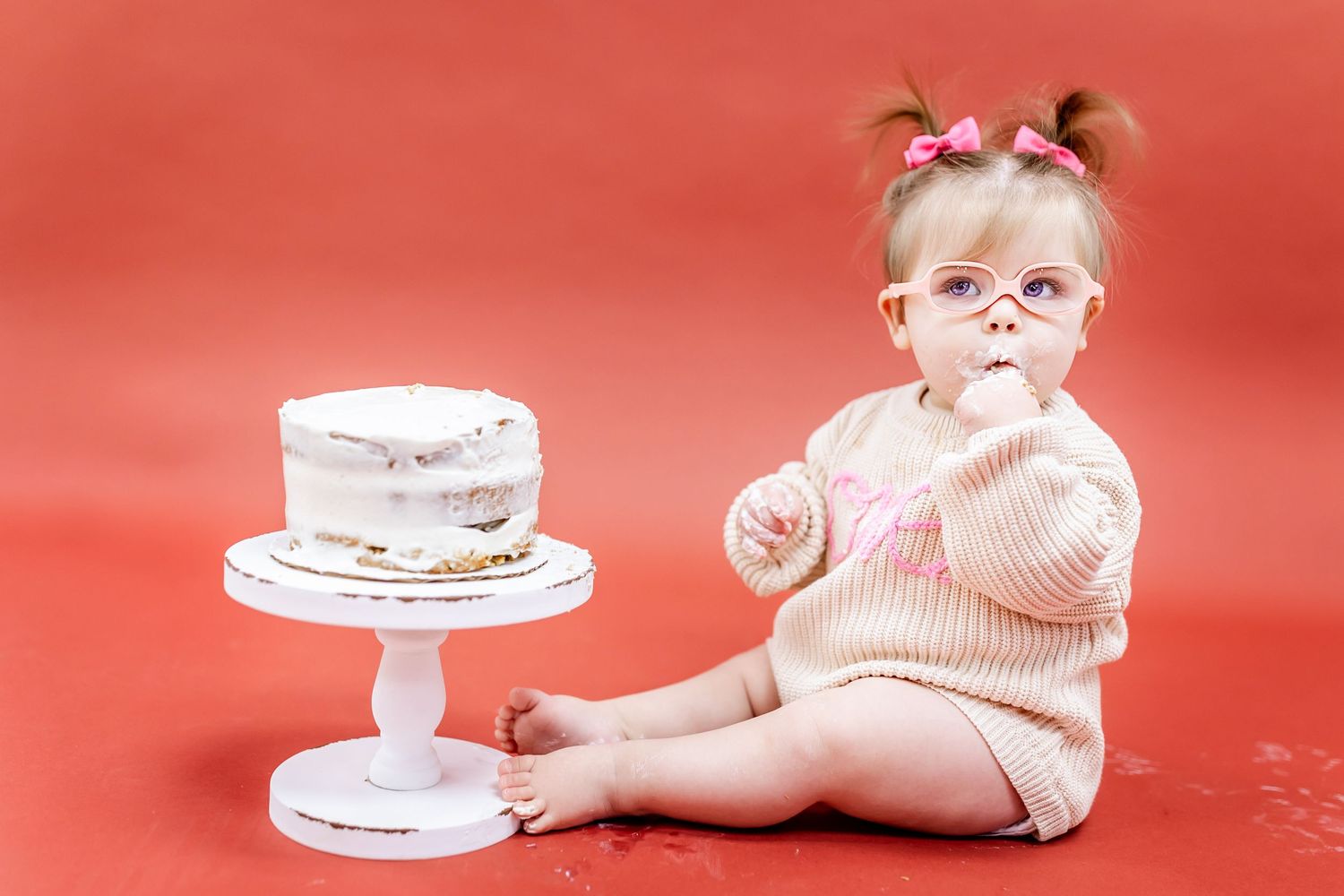 Baby with glasses sits next to white cake on stand against red background.