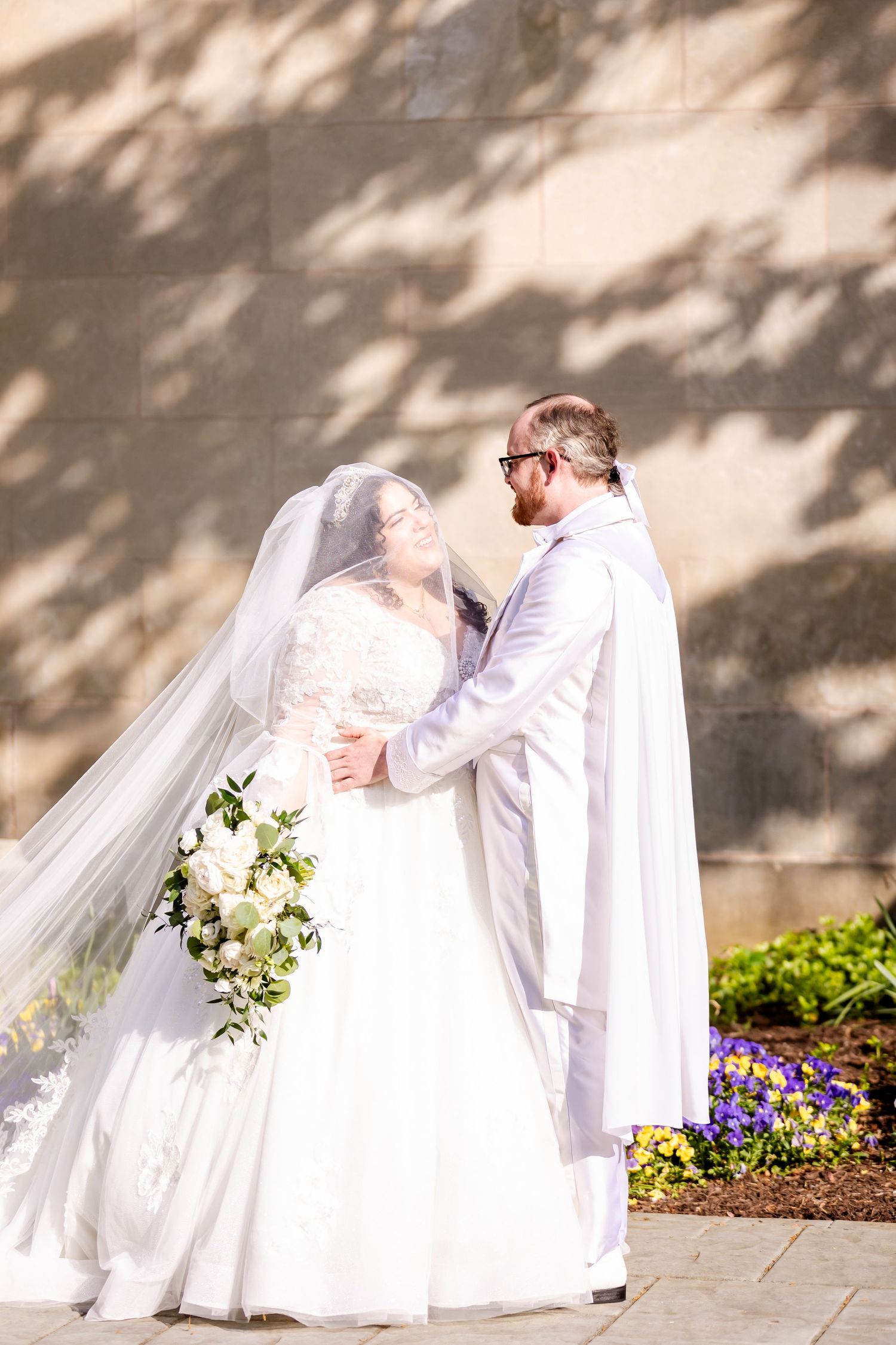 Intimate wedding portrait with couple embracing among shadows and purple flowers.