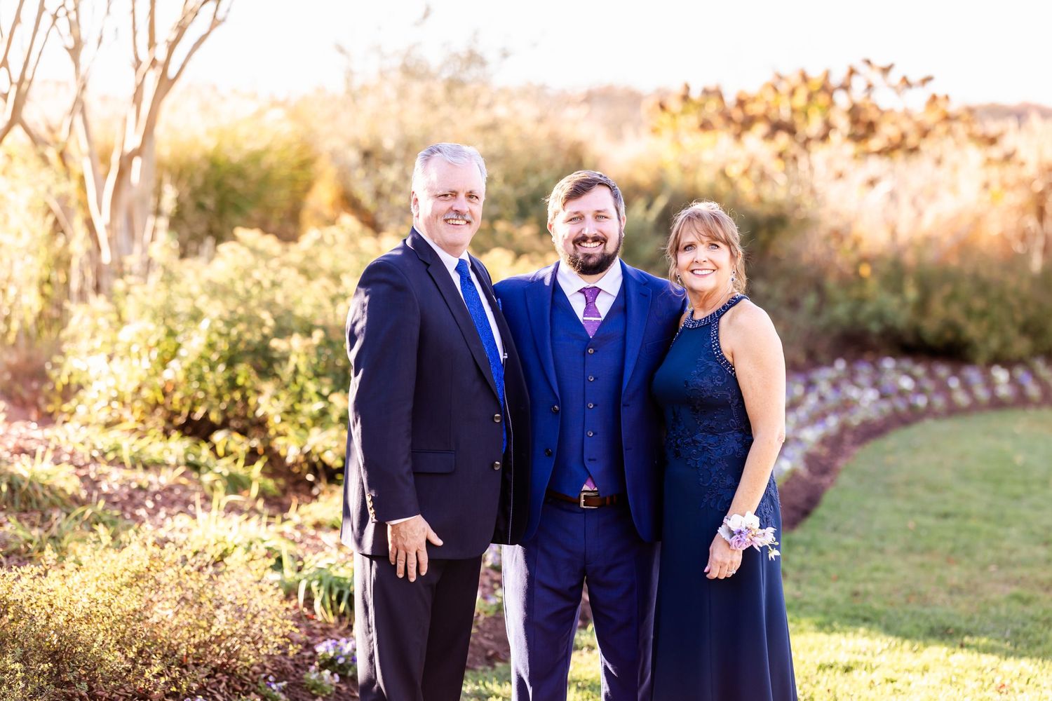 Wedding party members in coordinated navy formal wear pose together in a garden at golden hour.