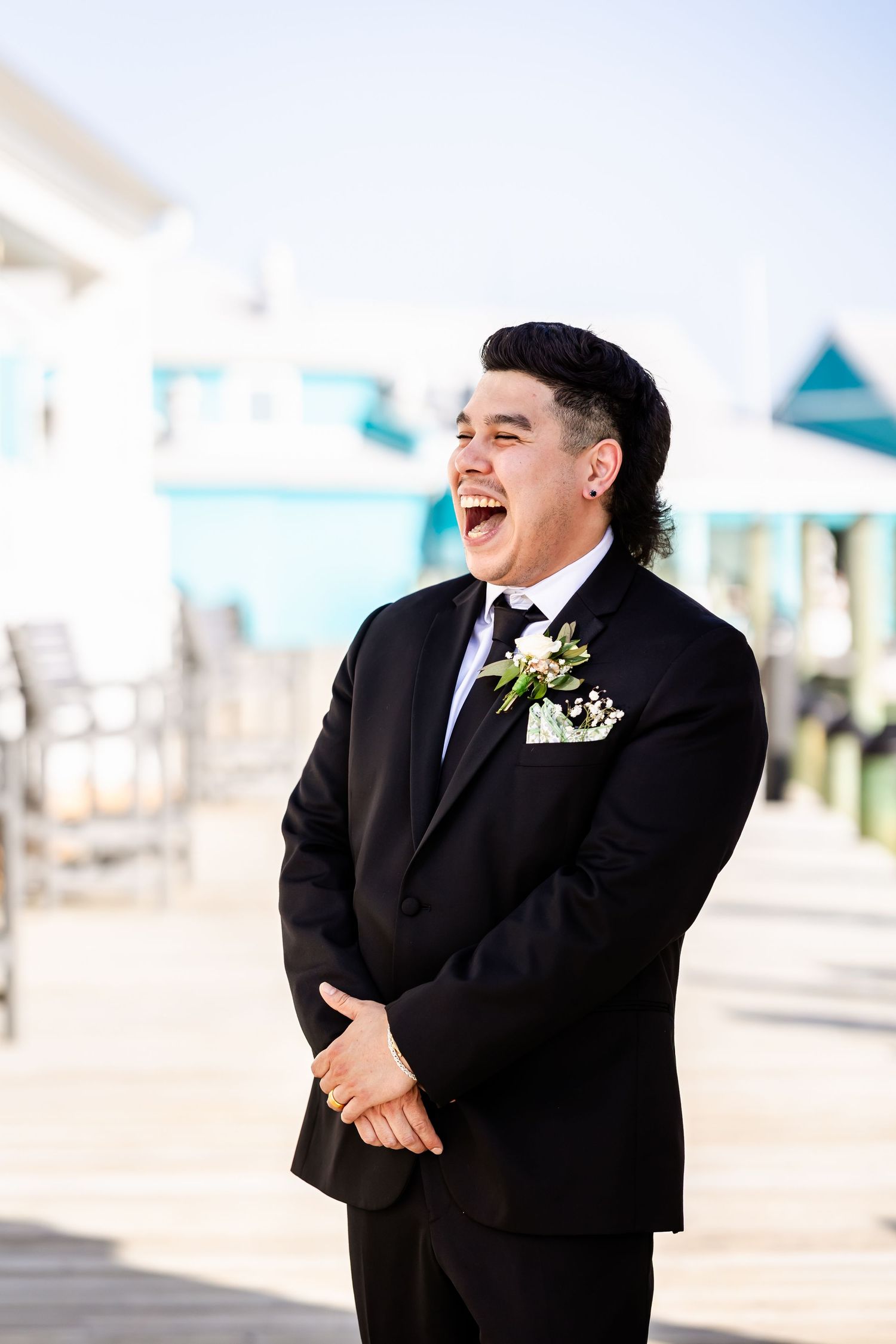 A groom in a classic black tuxedo and boutonniere laughs joyfully at a beachside wedding venue.