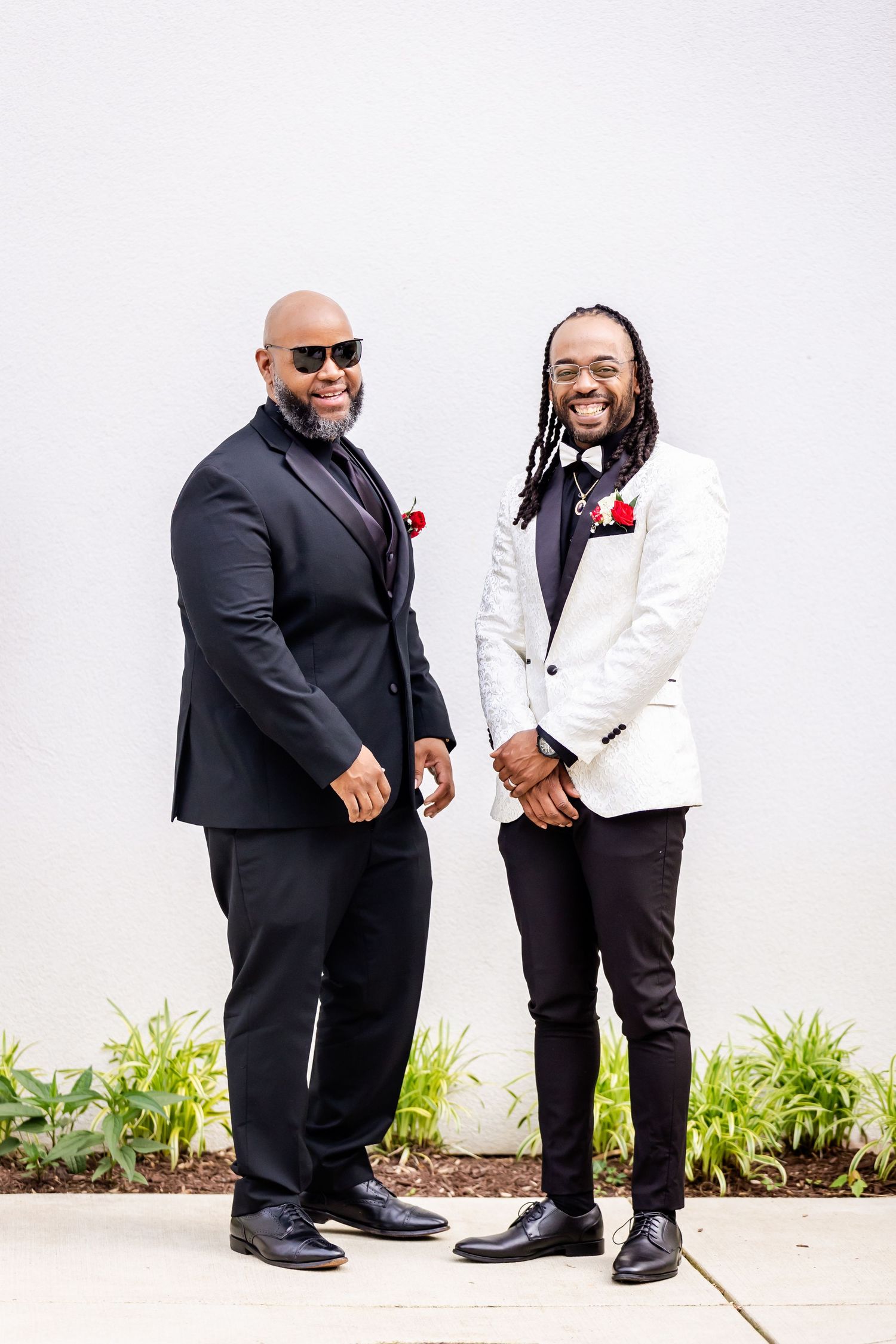 Two sharply dressed men in contrasting black and white suits pose against light wall.