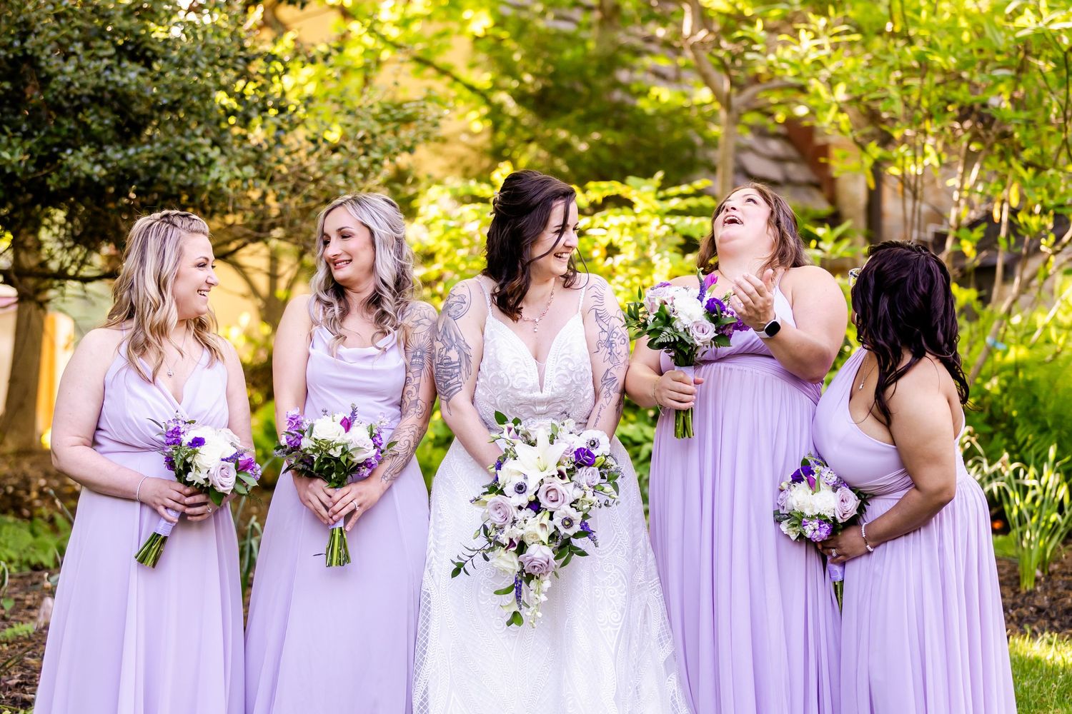 Wedding party wearing flowing lavender gowns holding floral bouquets in garden setting.