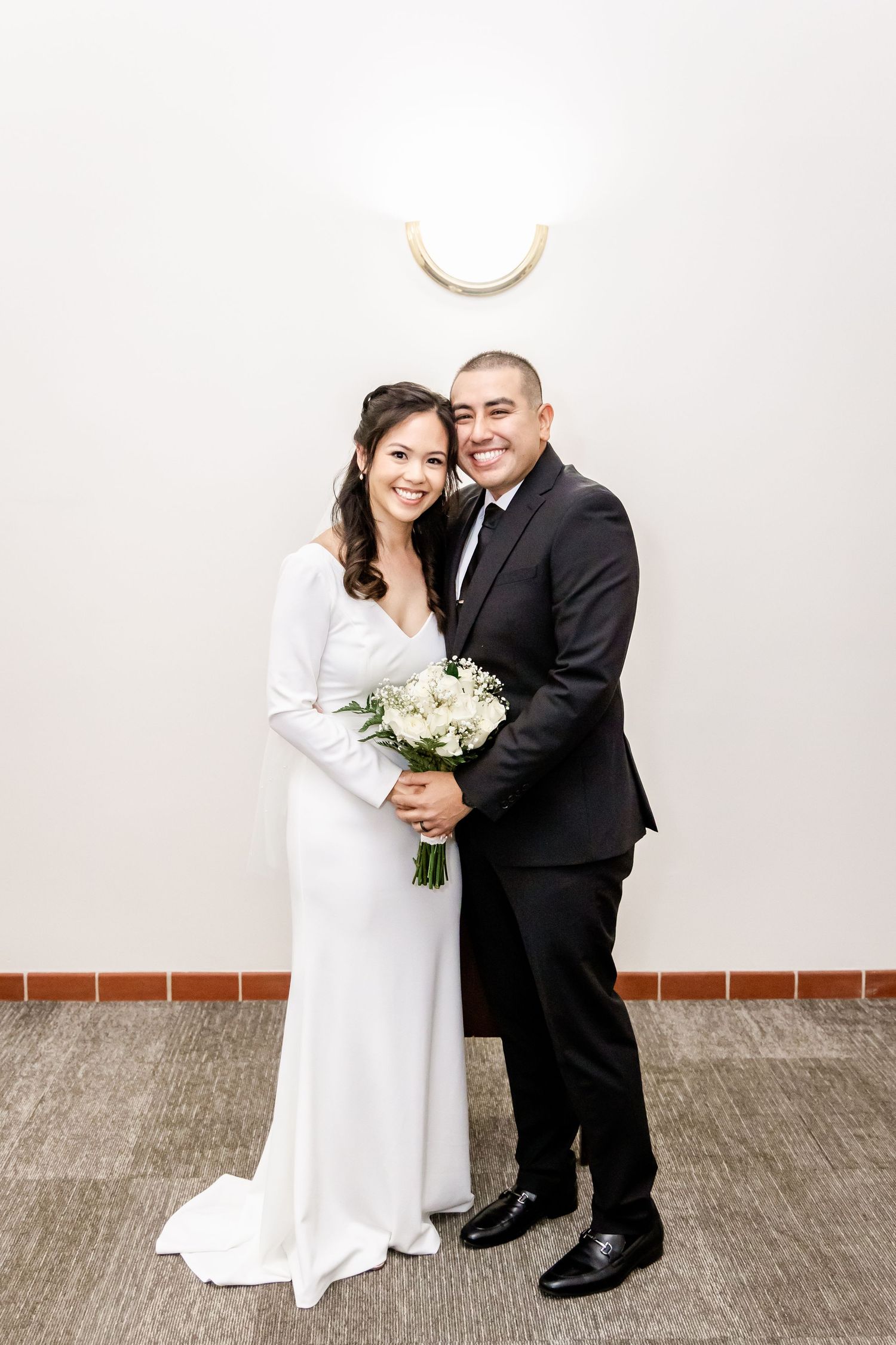Elegant couple poses with white bouquet during their wedding against minimalist backdrop.