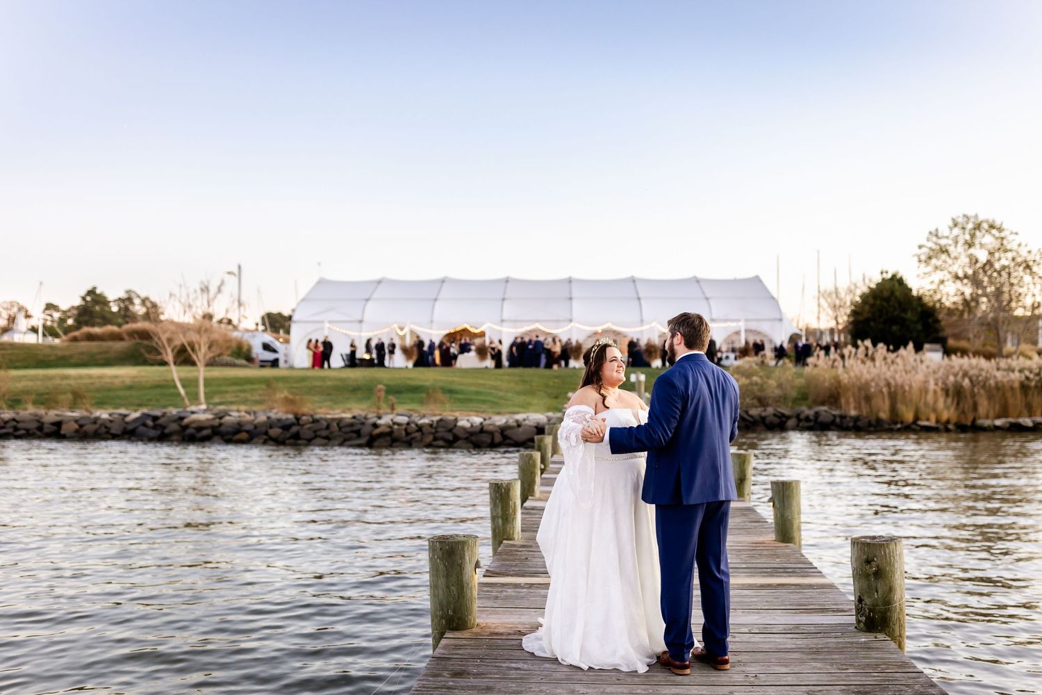 A wedding couple stands on a dock overlooking the water with a white tent reception in the background.