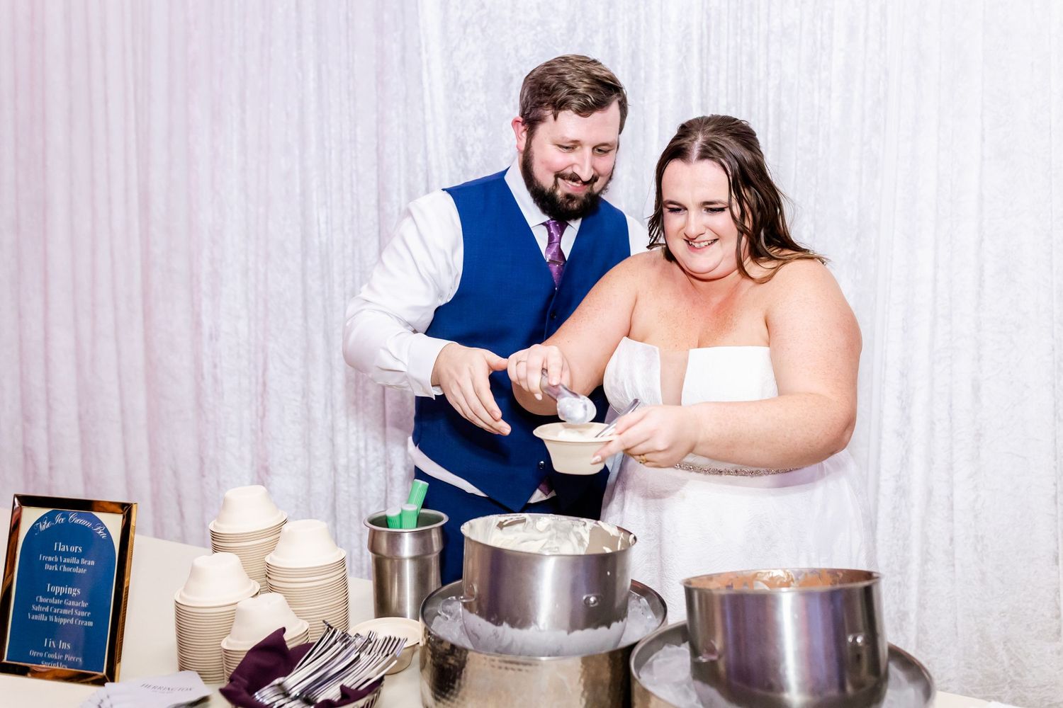 A couple serving food at a wedding buffet station with multiple silver chafing dishes and serving utensils.