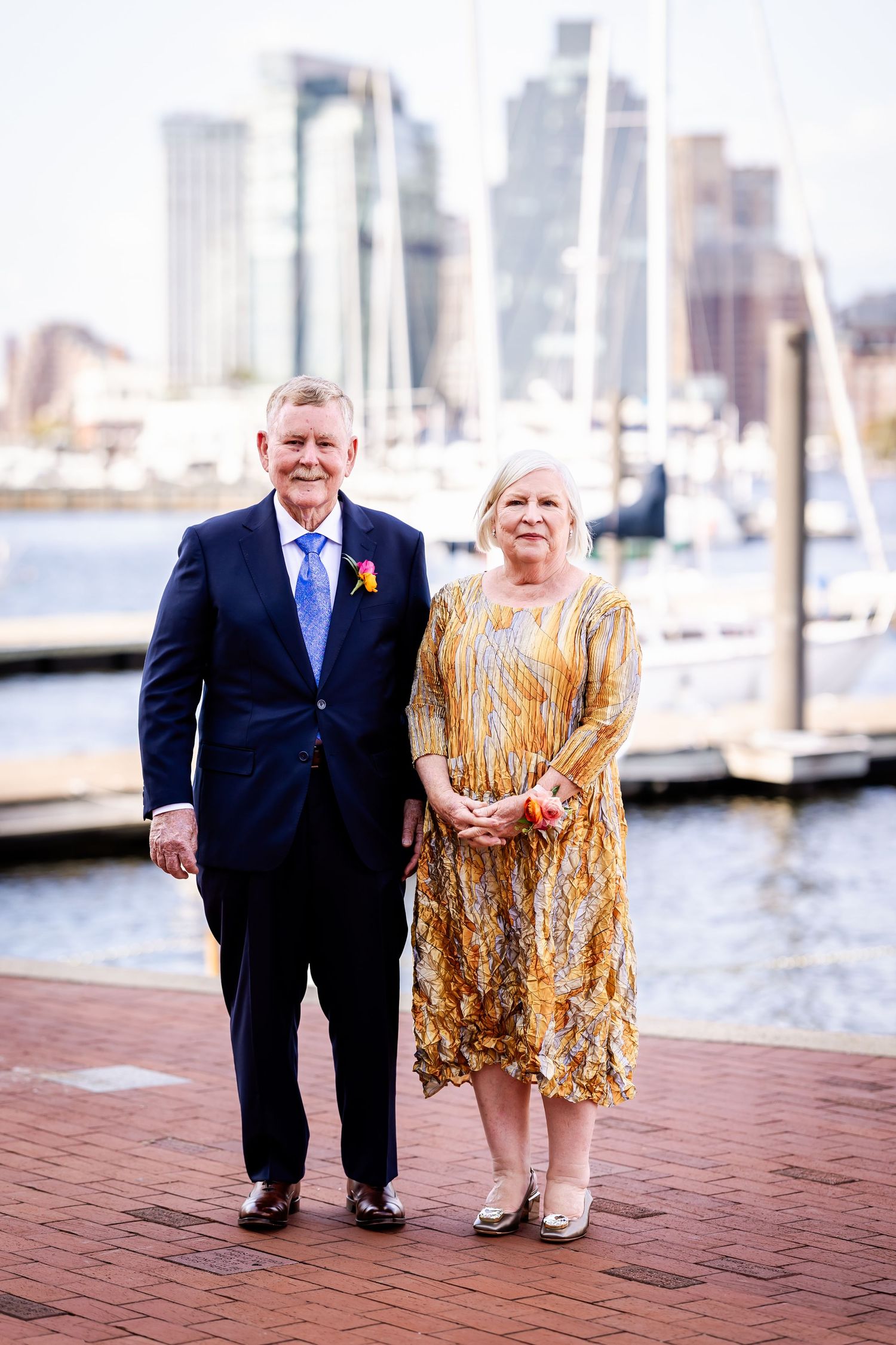Formal couple standing on brick waterfront with Baltimore harbor and skyline in background.