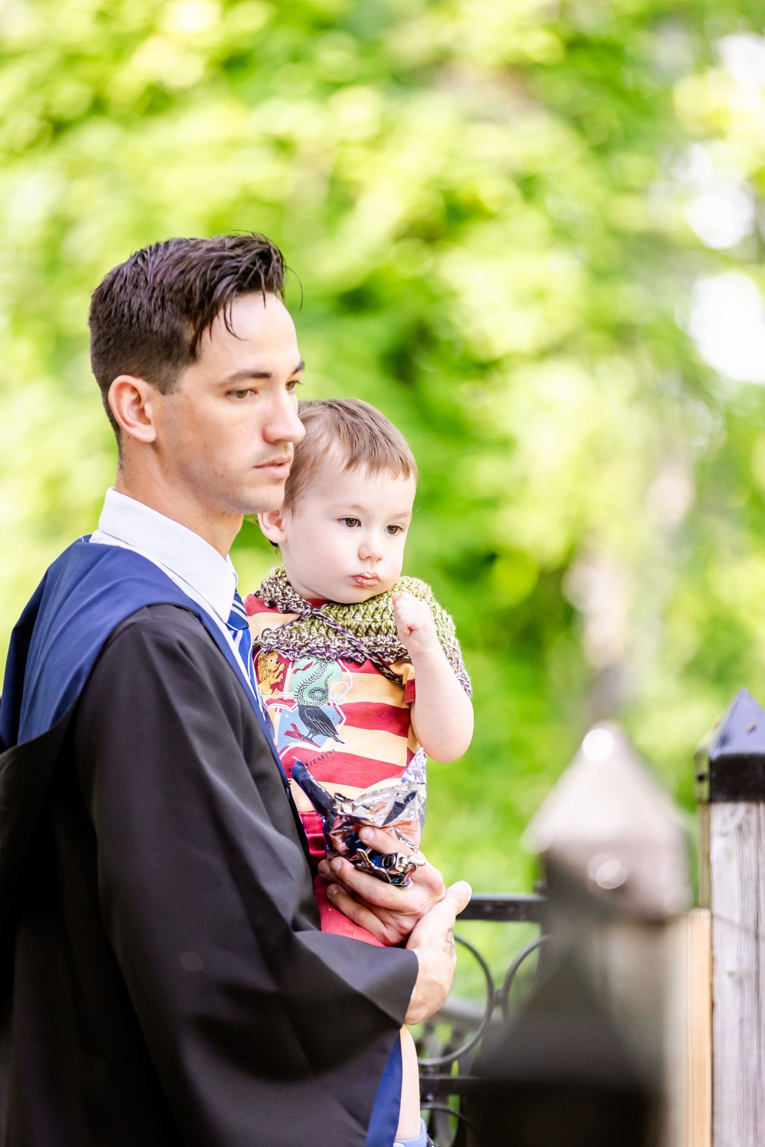 Parent in formal attire holding small child in patriotic shirt against green foliage backdrop.