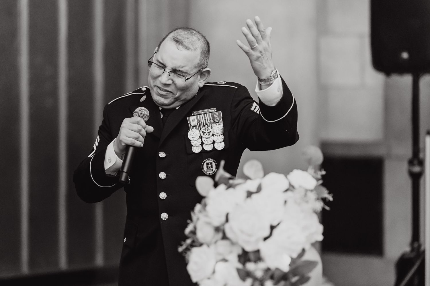 A uniformed servicemember speaks into a microphone while gesturing during a formal event with medals displayed on uniform.