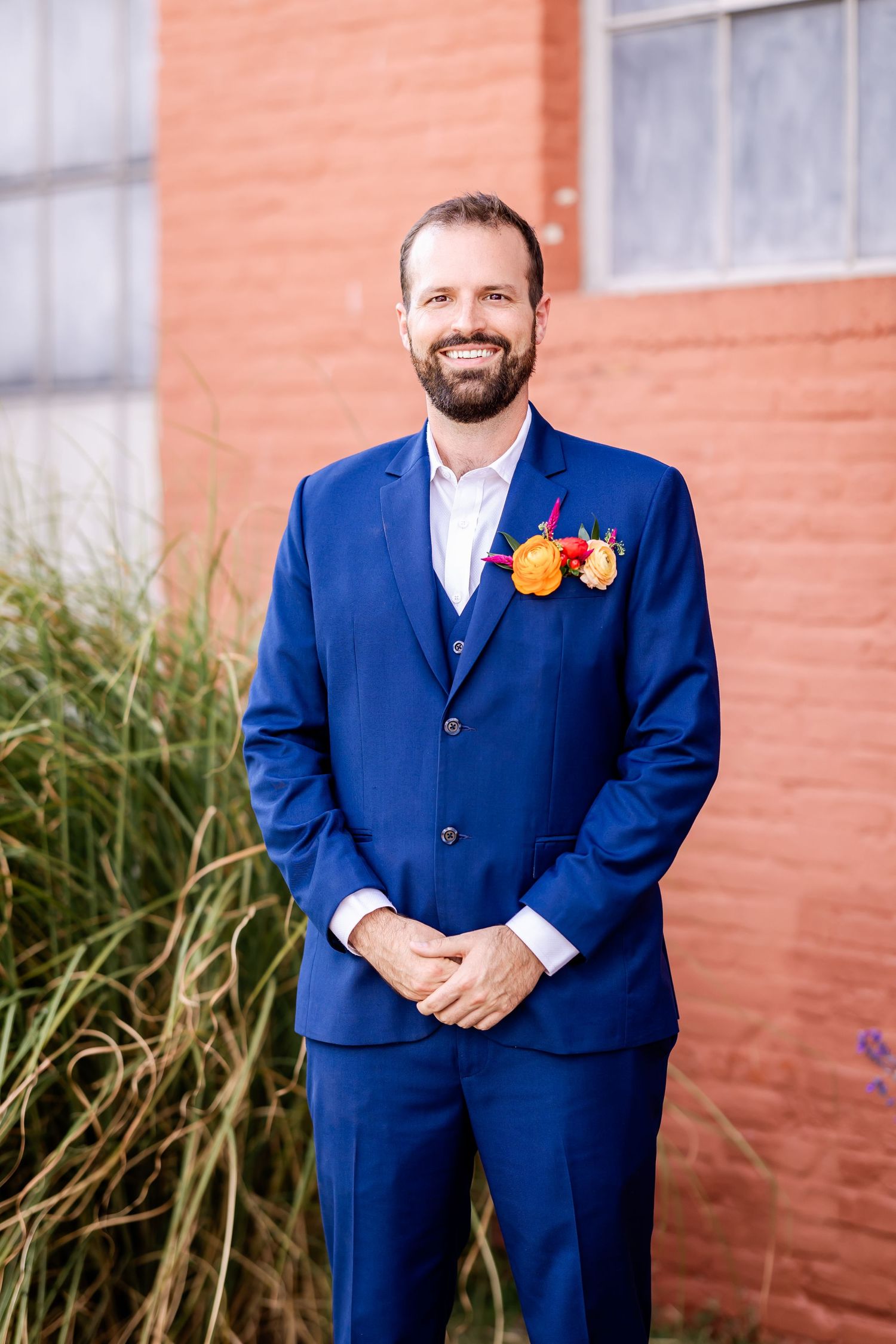A formal blue suit with orange and yellow floral boutonniere against a brick wall and ornamental grasses.