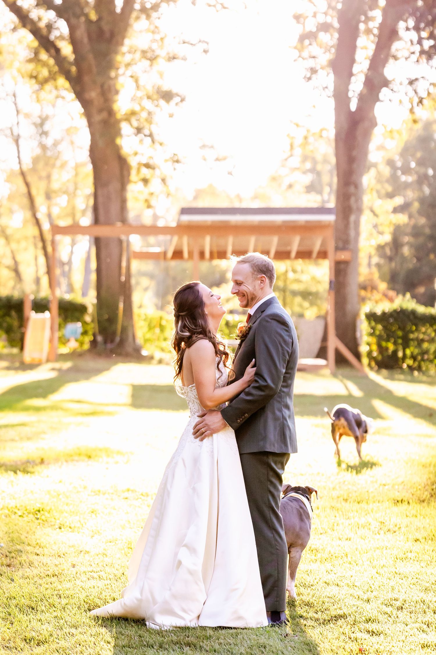 A romantic wedding couple embraces in a sunlit garden setting with golden autumn trees in the background.