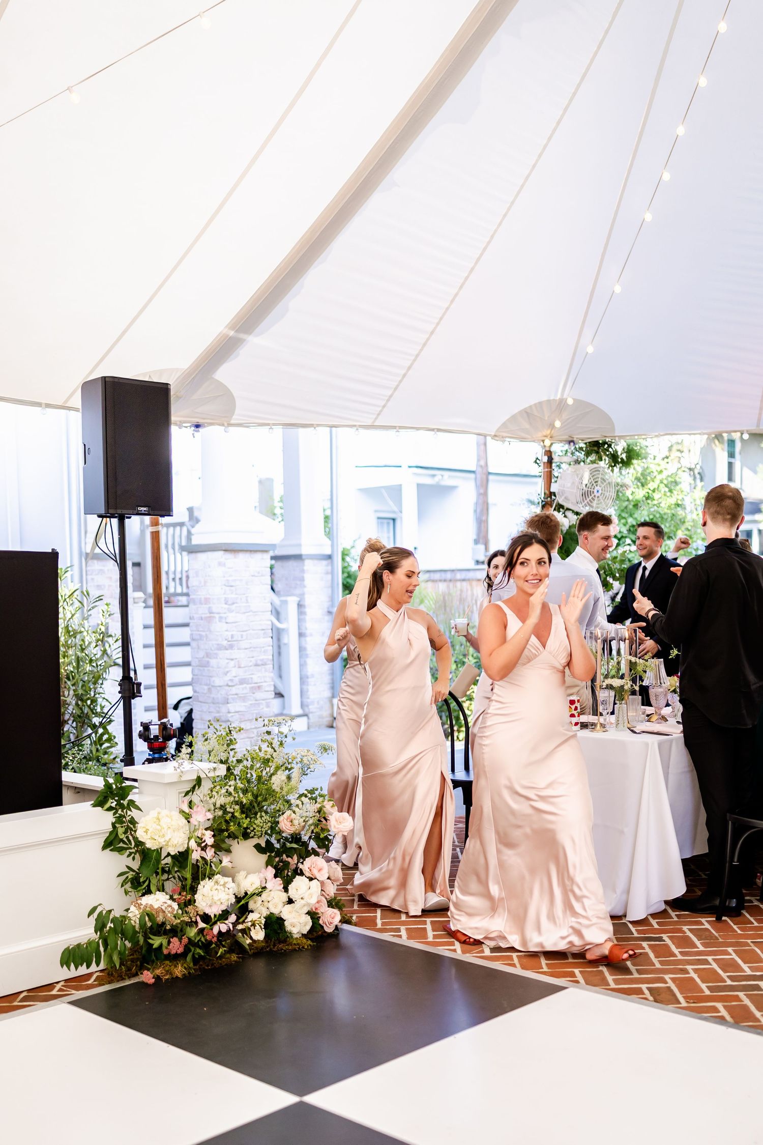 Bridesmaids in blush dresses gather near floral arrangements under an elegant white tent at a wedding reception.