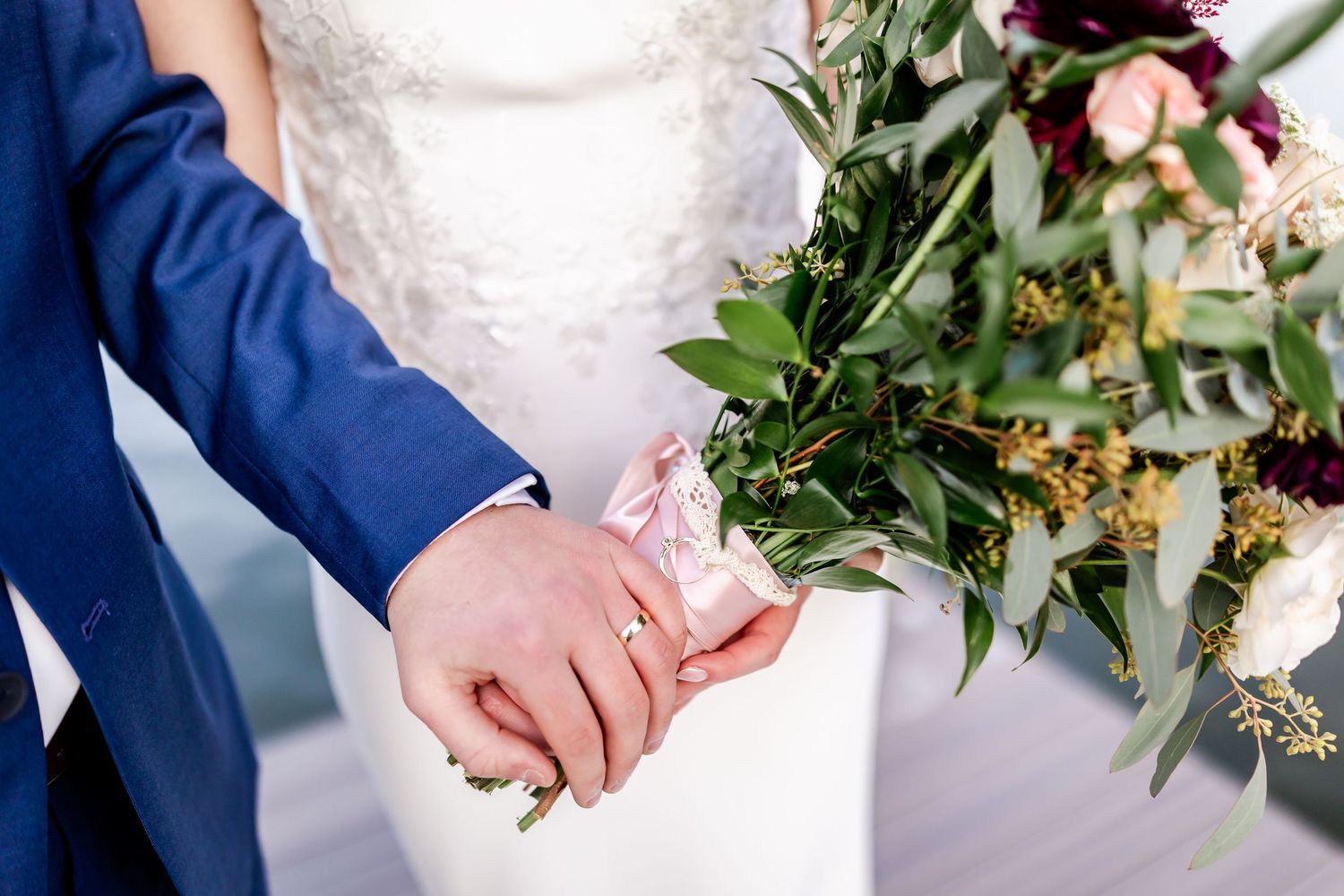 Close-up details of wedding attire showing white lace dress and navy suit with hands clasped together.