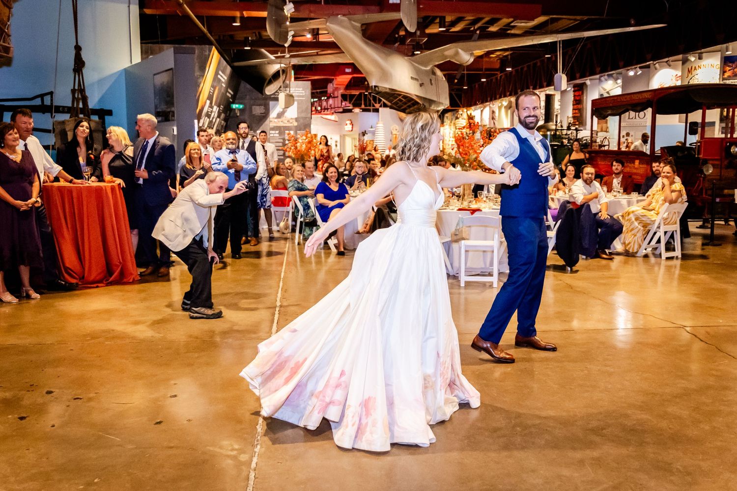 A couple performs their first dance in a rustic venue with guests watching from decorated tables.