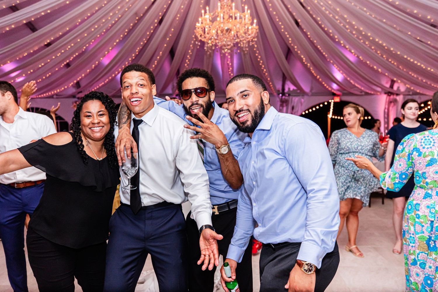 People dance and celebrate at an elegant event under a tent decorated with fabric draping and a crystal chandelier.