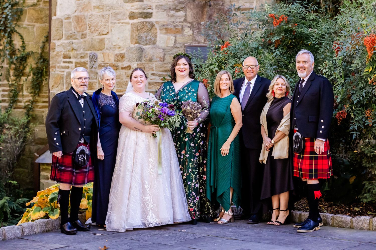 Wedding group photo featuring Scottish kilts and formal attire against stone church backdrop with autumn decorations.