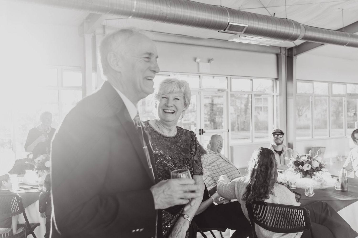 A candid black and white photograph capturing a joyful shared moment at an indoor reception or gathering.