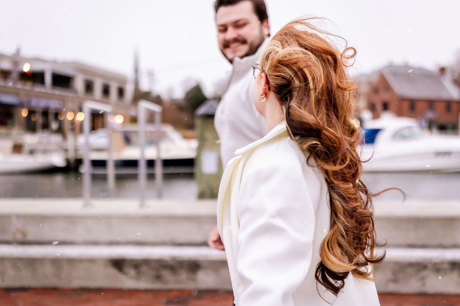 A couple in white sweaters shares a joyful moment together by a marina with boats in the background.