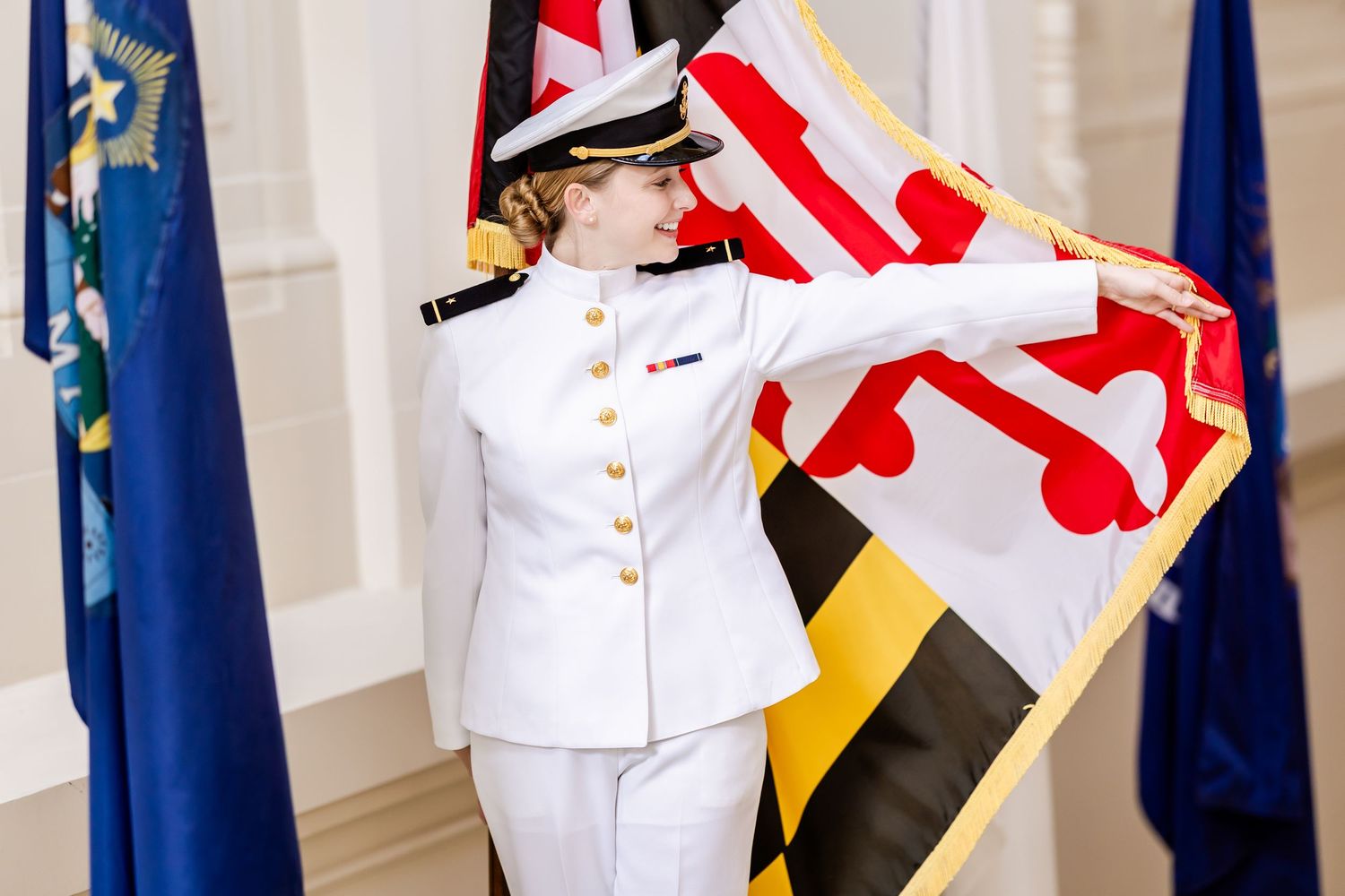 Naval officer in white dress uniform stands at attention with state flag display.