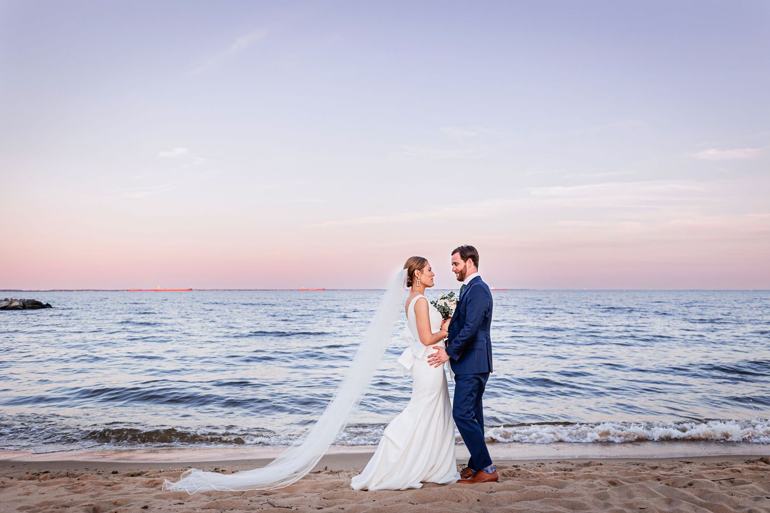 A lovely couple standing on a beach at sunset embracing, with bride wearing a long flowing gown and veil.