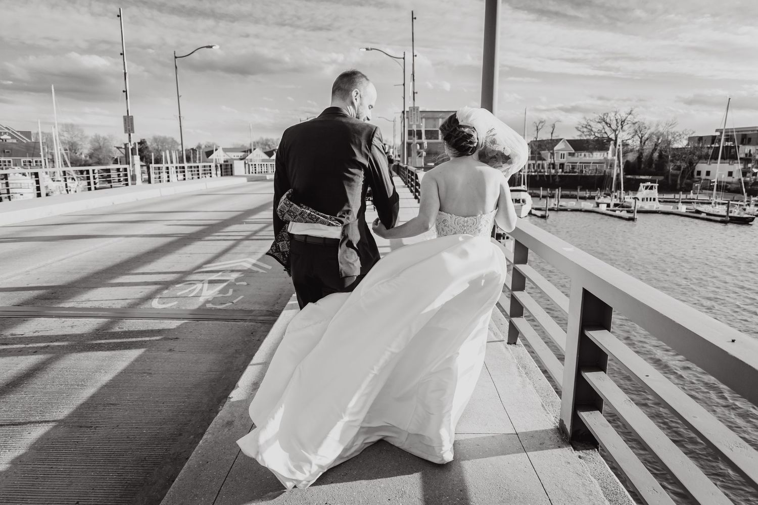 Black and white photo of couple walking on boardwalk with bride's dress flowing in the wind.