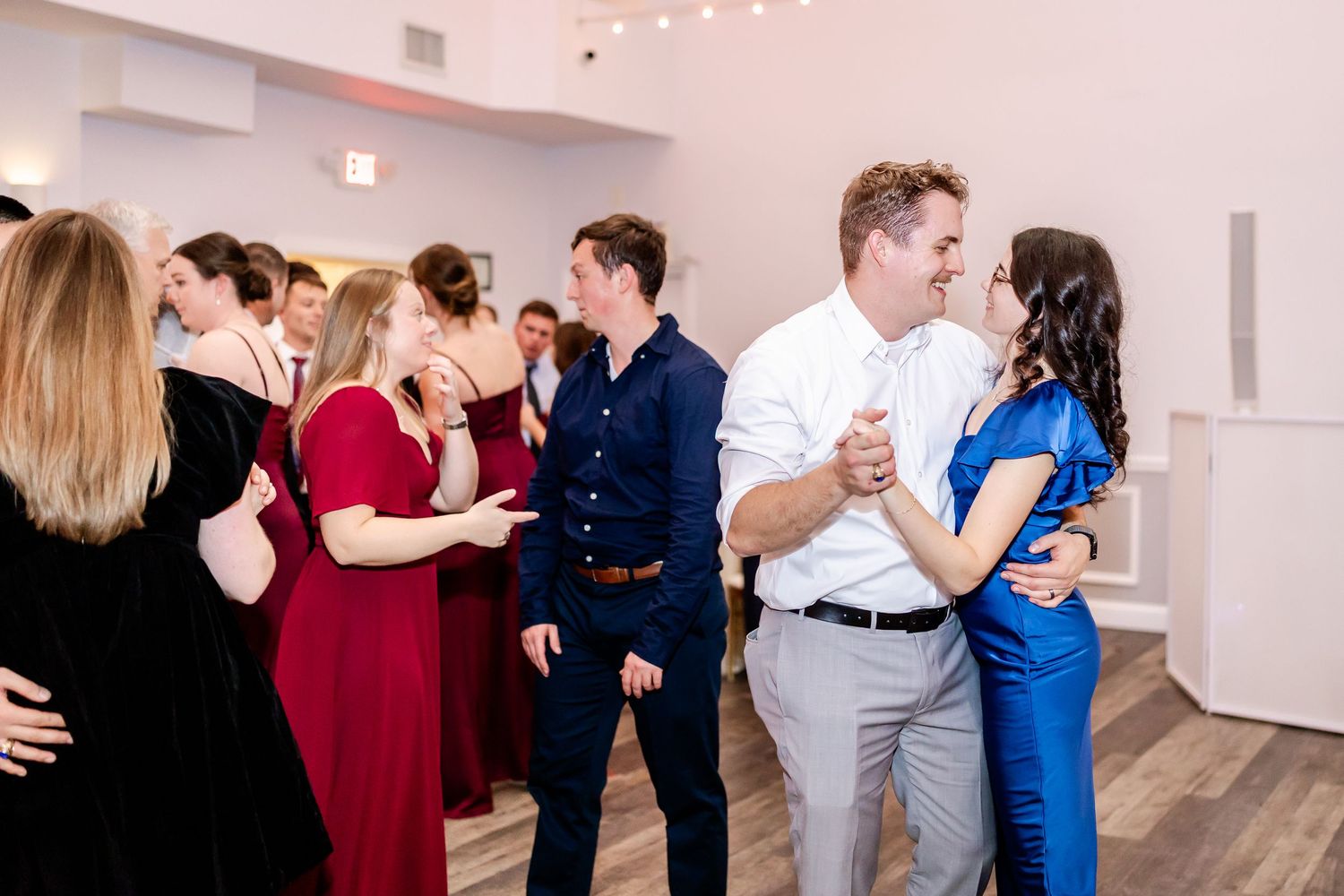 Wedding guests dancing and mingling at an elegant reception with white walls and hardwood floors.