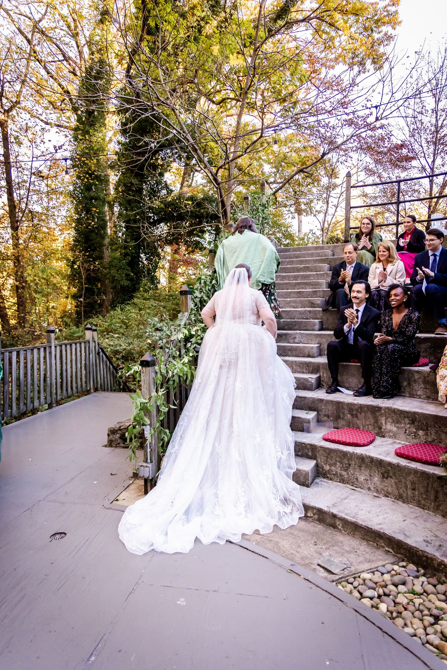 Bride in flowing white gown ascending stone steps outdoors while guests watch from above.