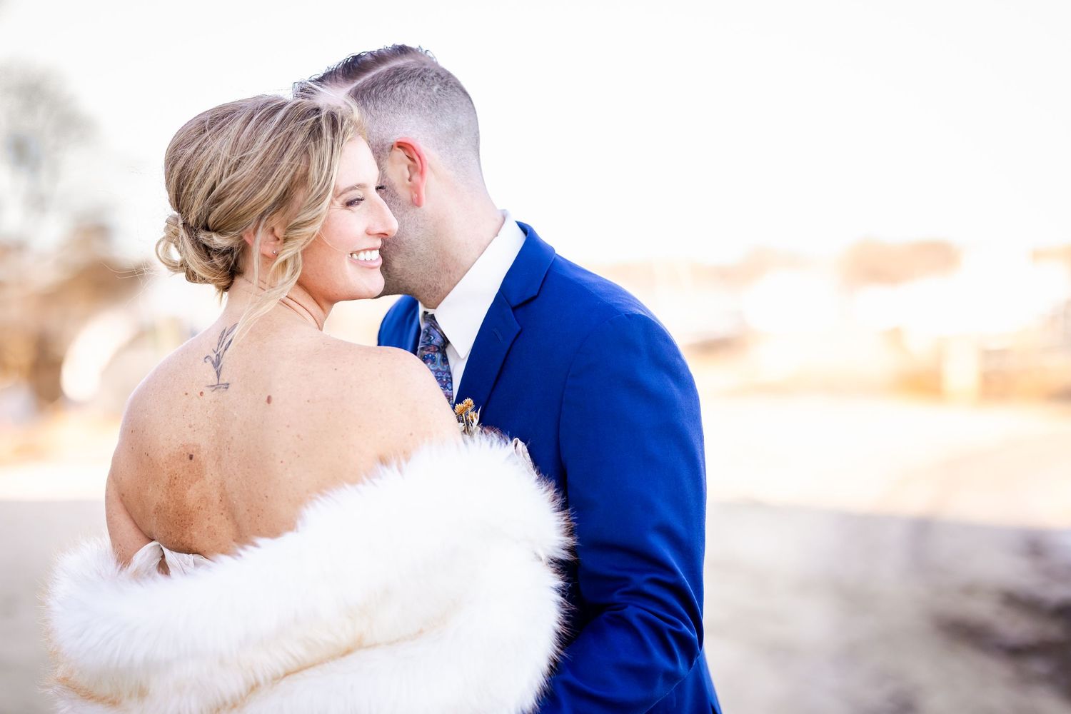 A romantic wedding couple embraces outdoors with the bride wearing white fur and the groom in a royal blue suit.