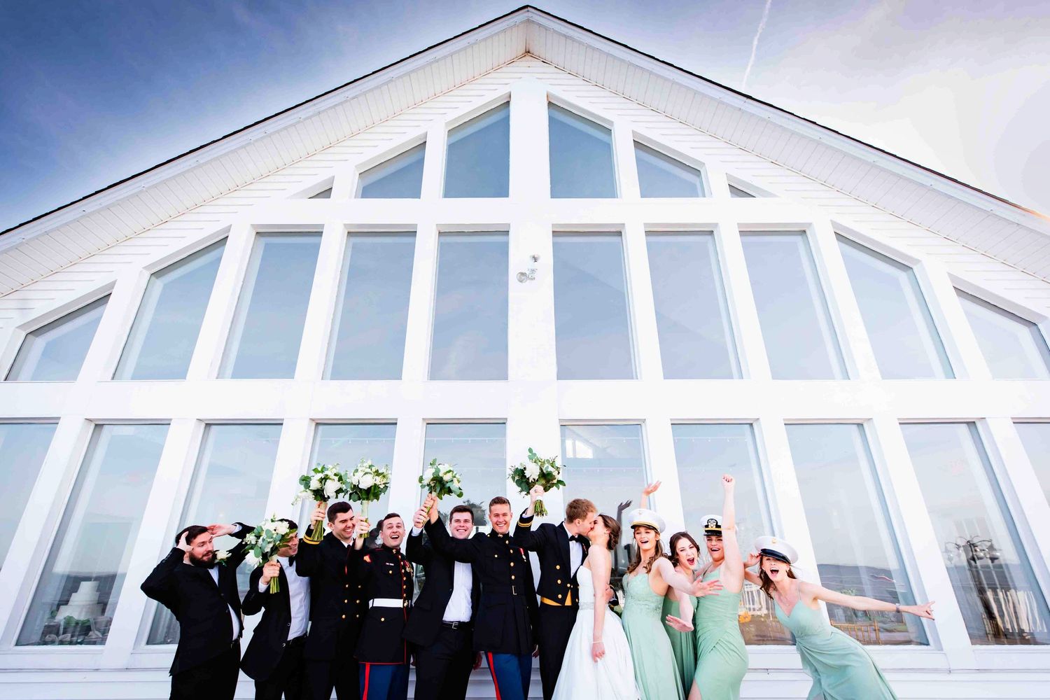 Wedding party poses together in front of a modern white chapel with large triangular windows and bright lighting.