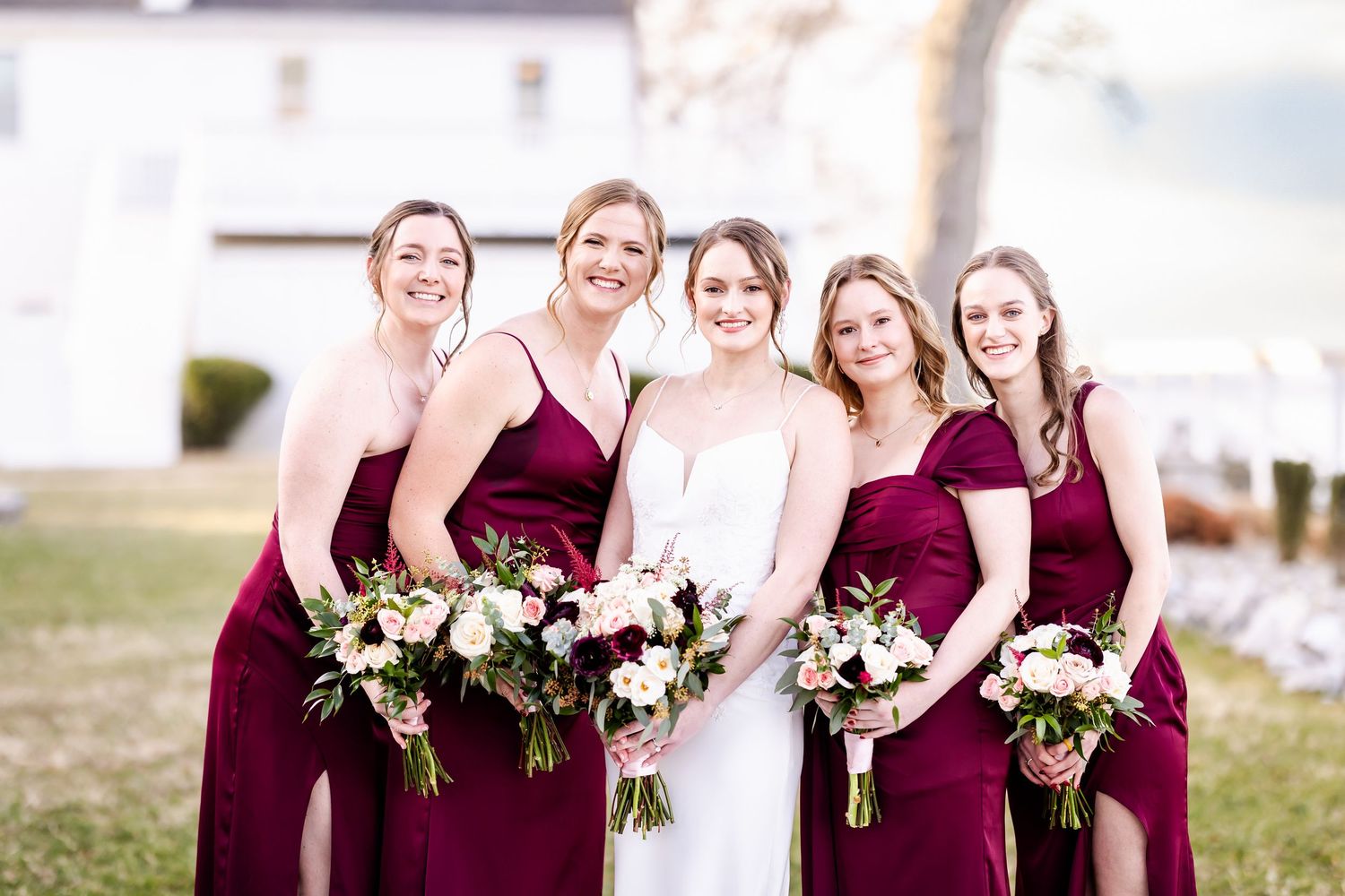 Wedding party poses outdoors holding white and burgundy floral bouquets in coordinated burgundy dresses.