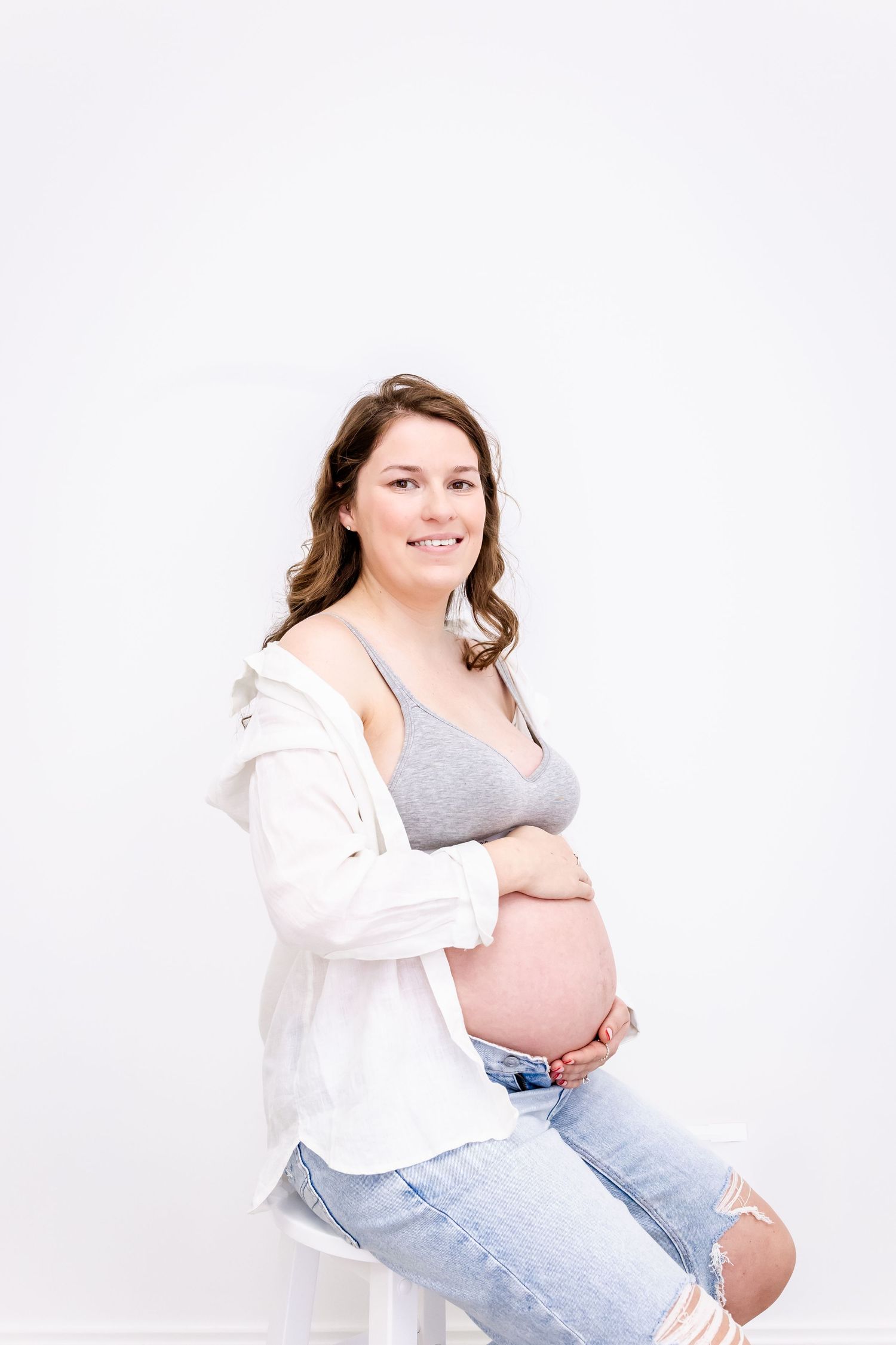 Expectant mother in casual attire sitting on white stool wearing gray tank top, white cardigan and distressed jeans.