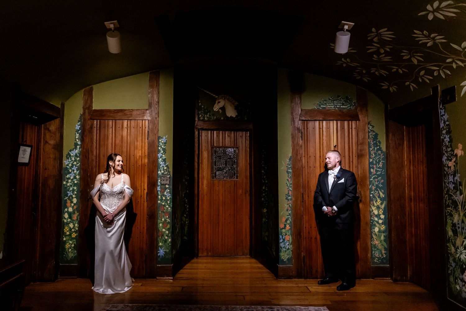 A couple in wedding attire poses in a dimly lit church hallway with wooden paneling and green floral decor.
