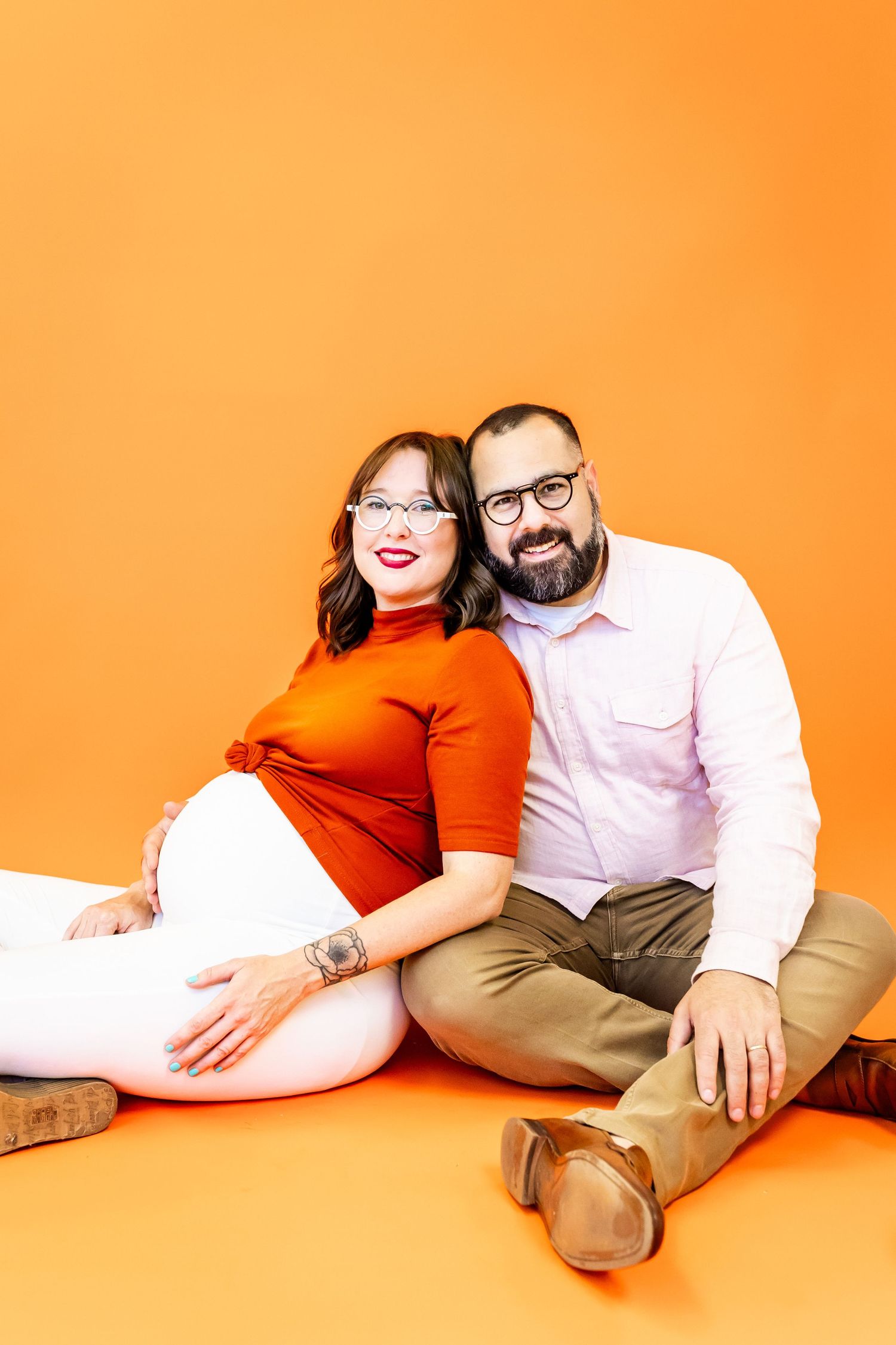Couple lounging together on orange background in casual attire.