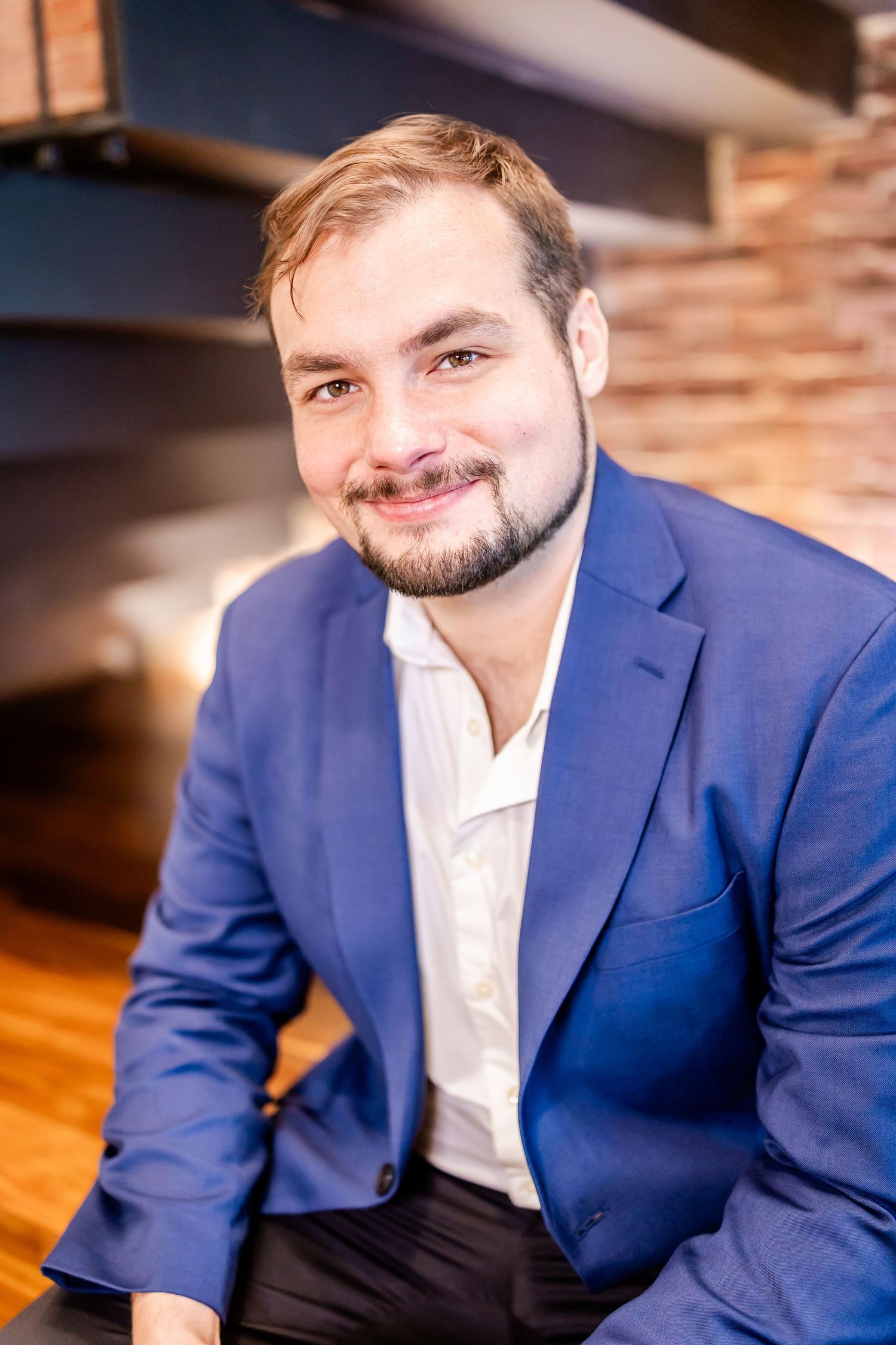 Professional headshot of person in navy blazer and white shirt against wooden wall background.